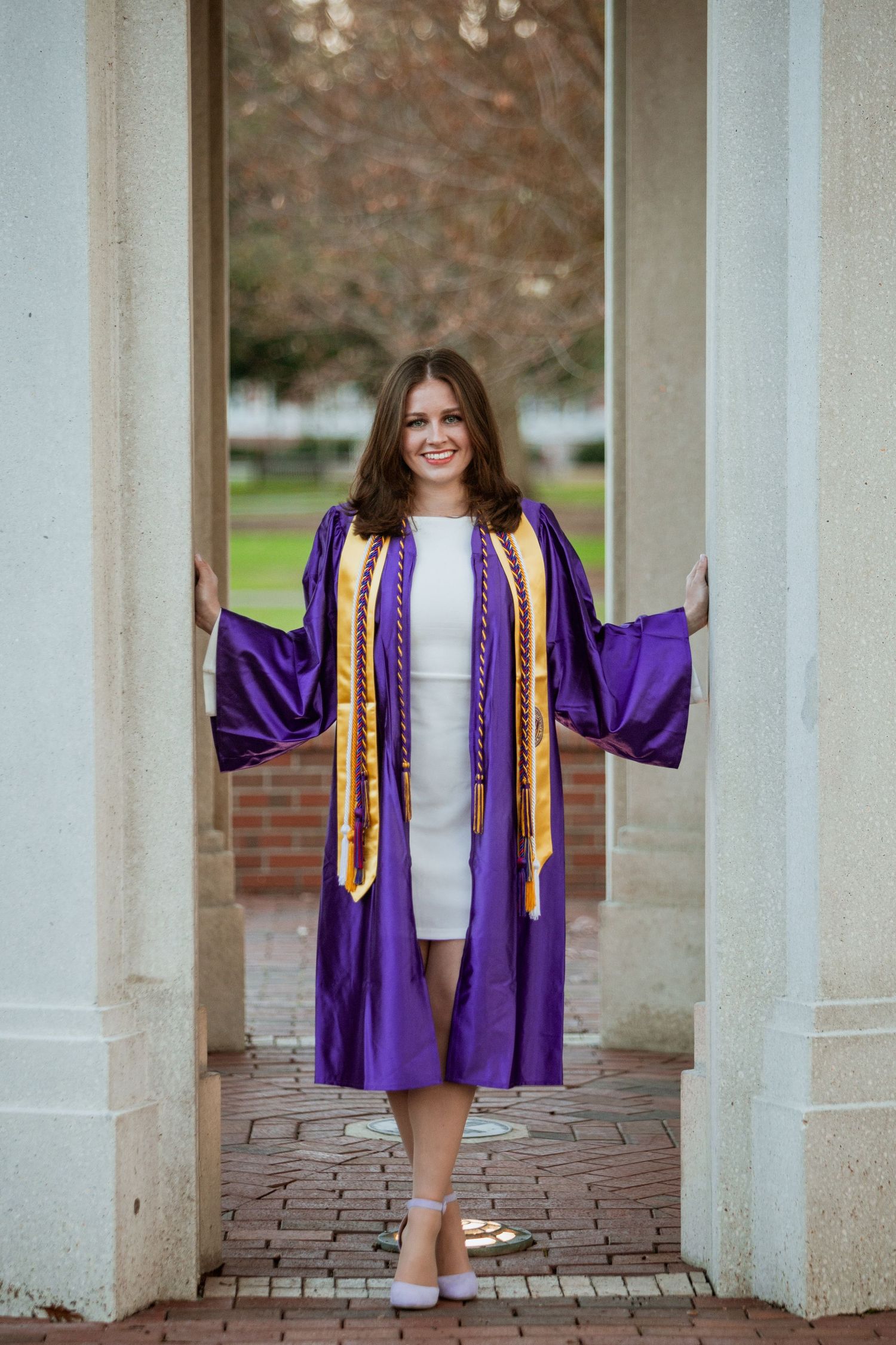 ECU graduate in purple graduation gown with gold stole stands between white campus columns smiling.