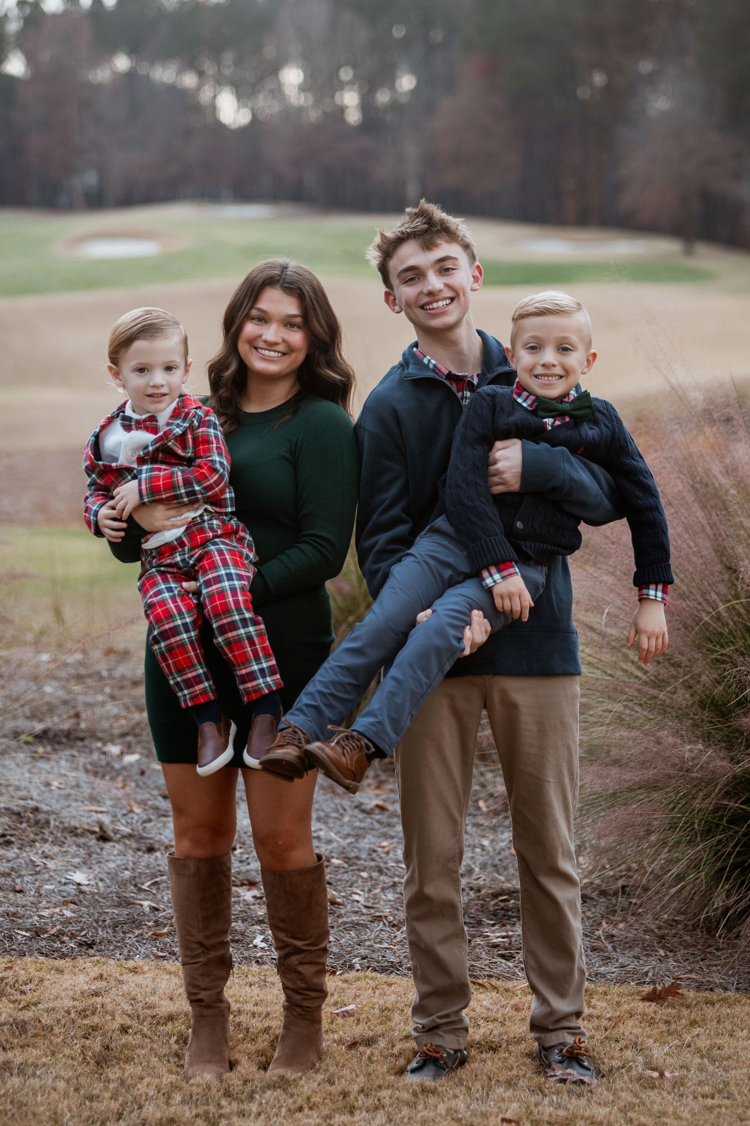 Four siblings pose outdoors, two teens holding two young boys in festive holiday outfits near a golf course.