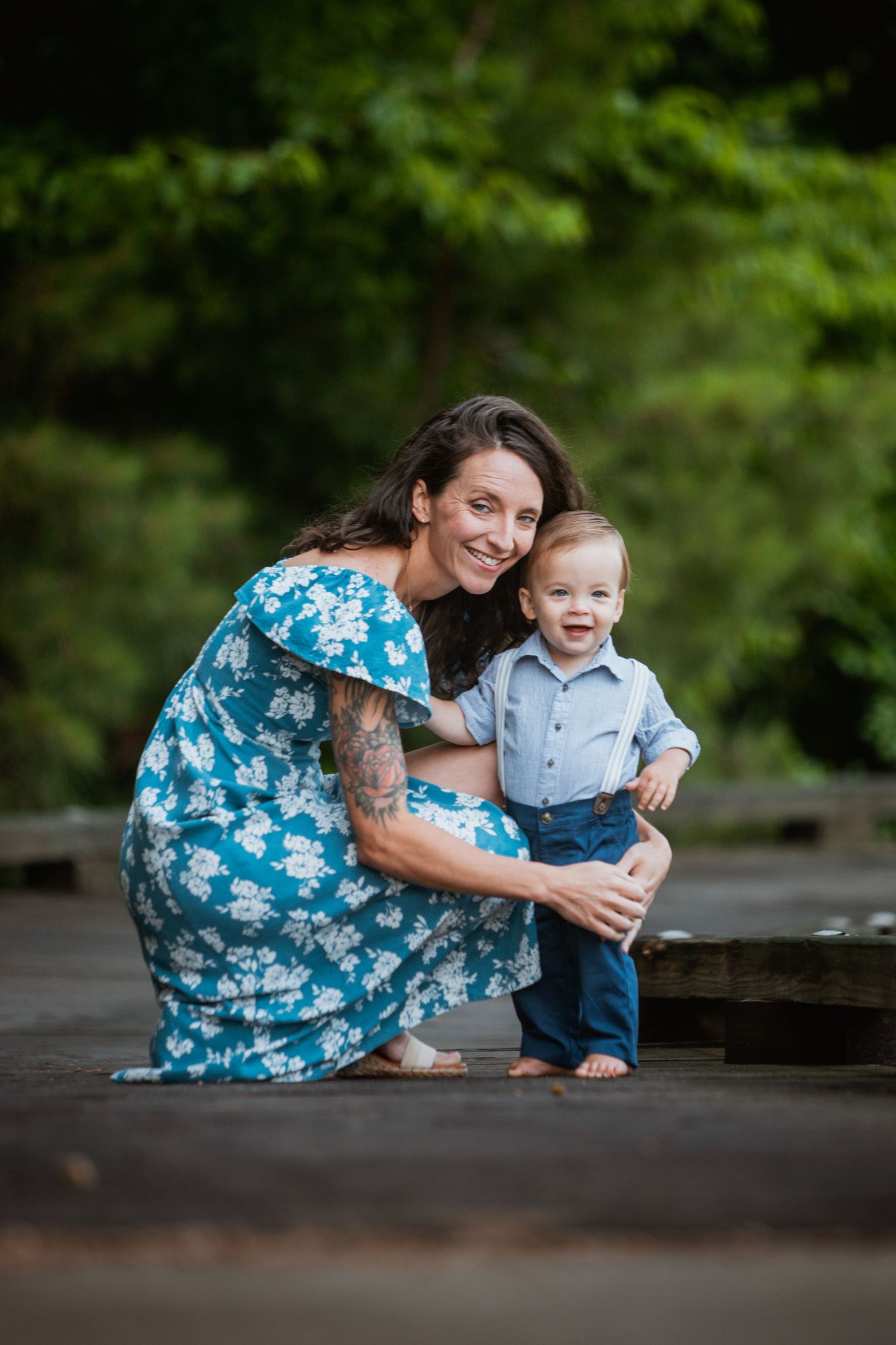 Smiling mother in blue floral dress crouching beside her happy toddler son on a wooden deck outdoors.