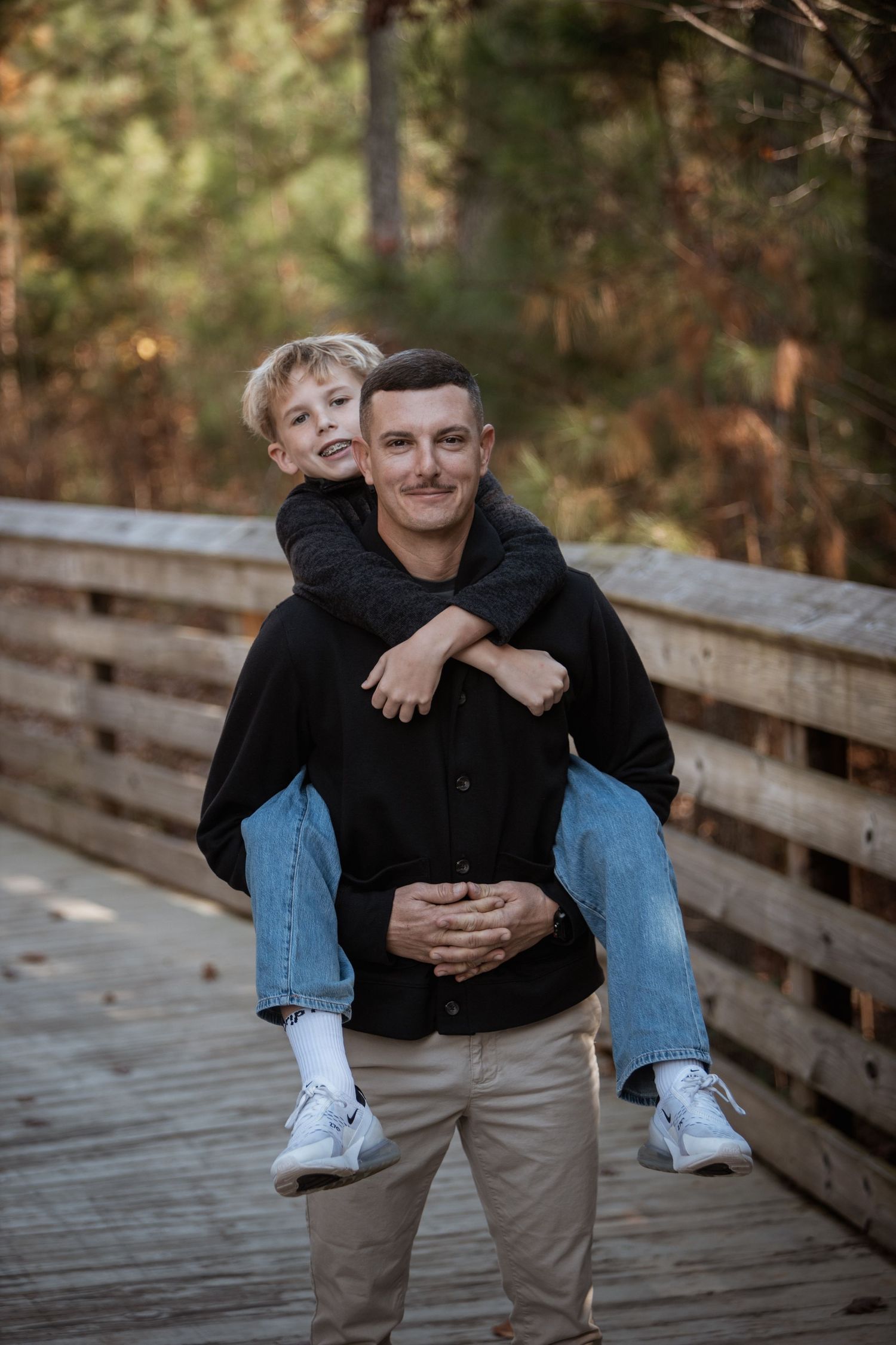 Man giving a laughing young boy a piggyback ride on a wooden bridge outdoors.