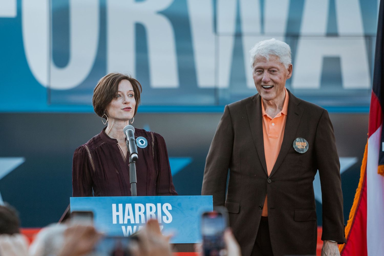 Two speakers at a Harris Walz campaign rally stand at a podium on stage before a crowd of supporters.