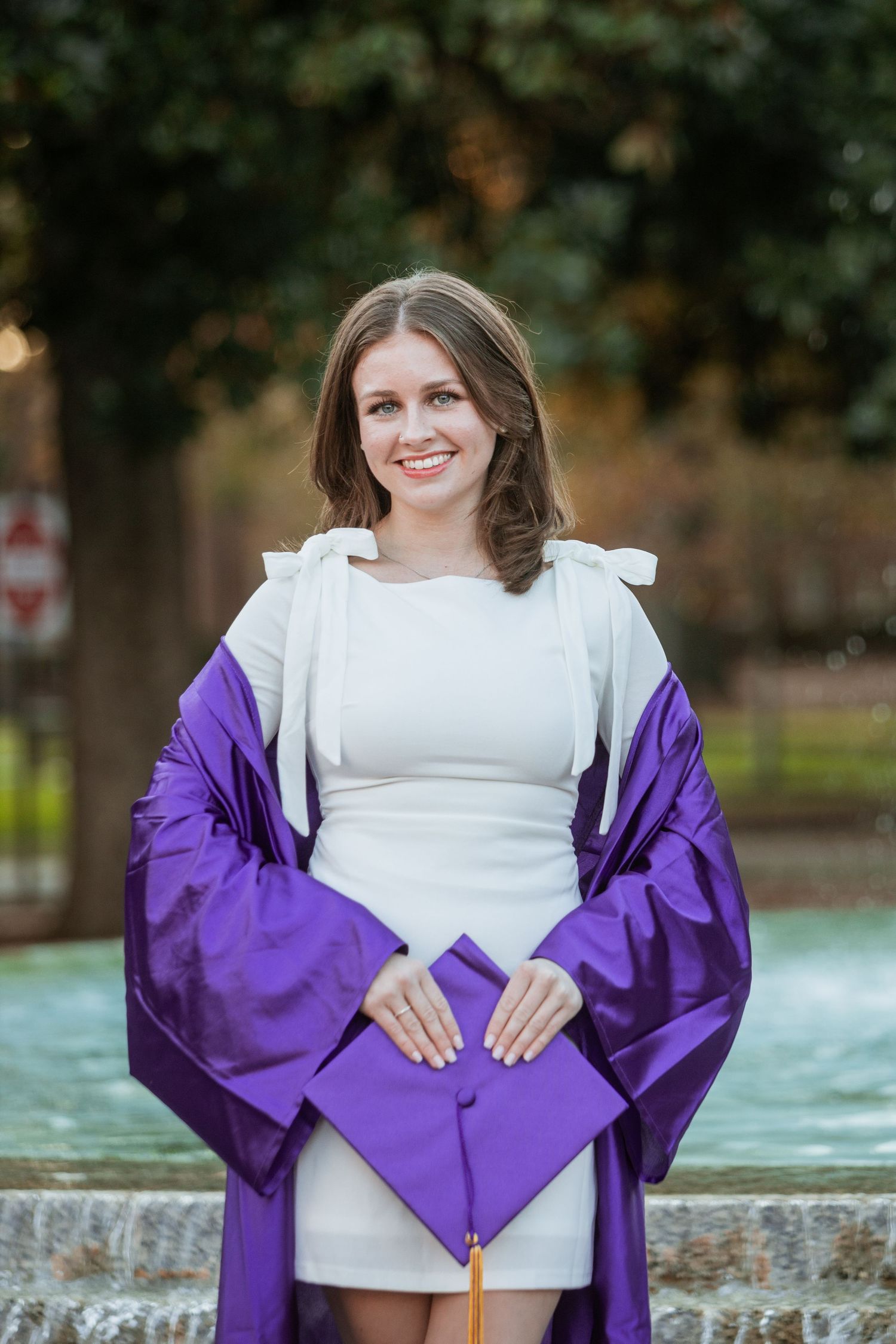 Happy graduate in purple cap and gown over white dress poses by campus fountain smiling.