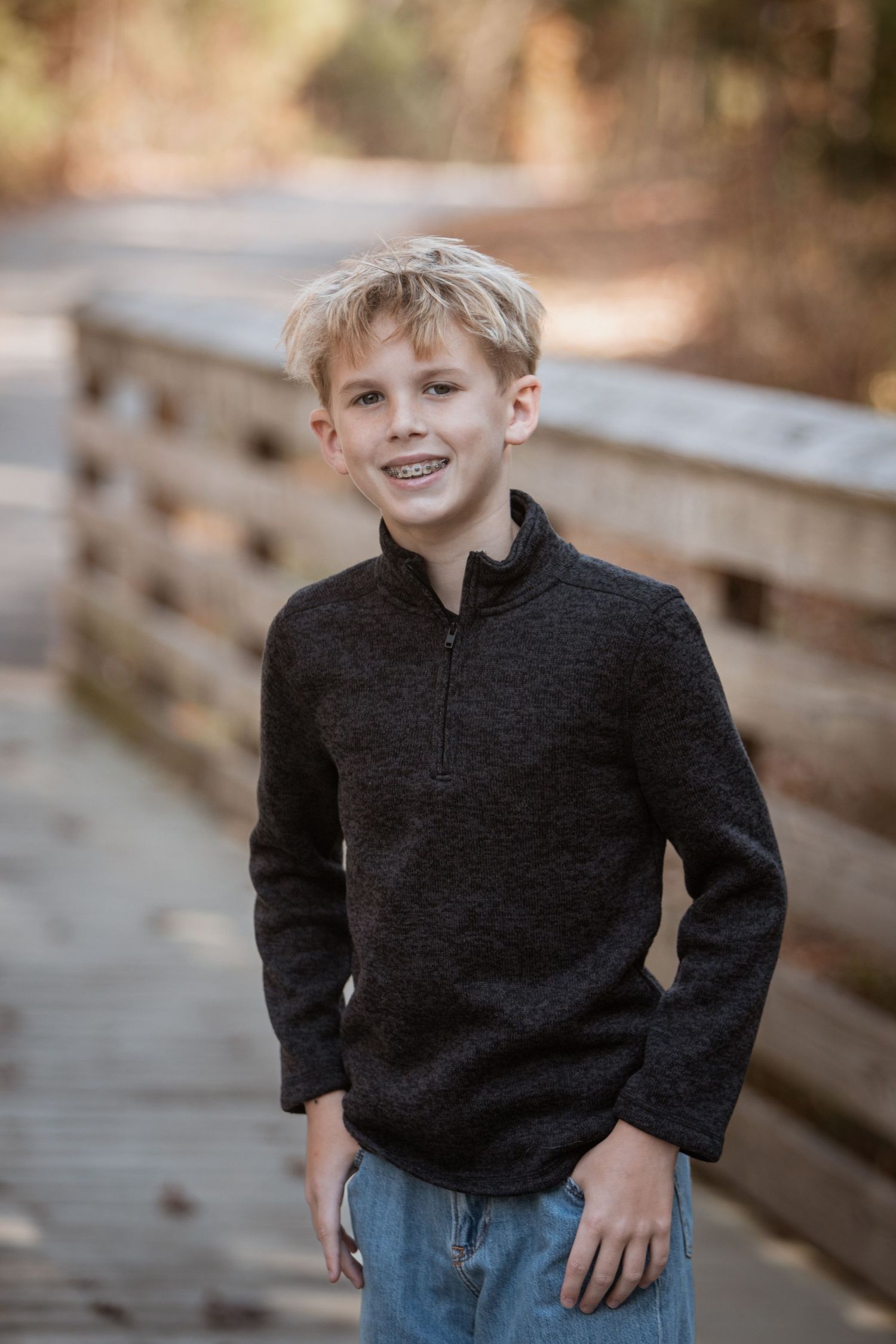 Smiling blonde boy in dark zip pullover standing on a wooden bridge in autumn.
