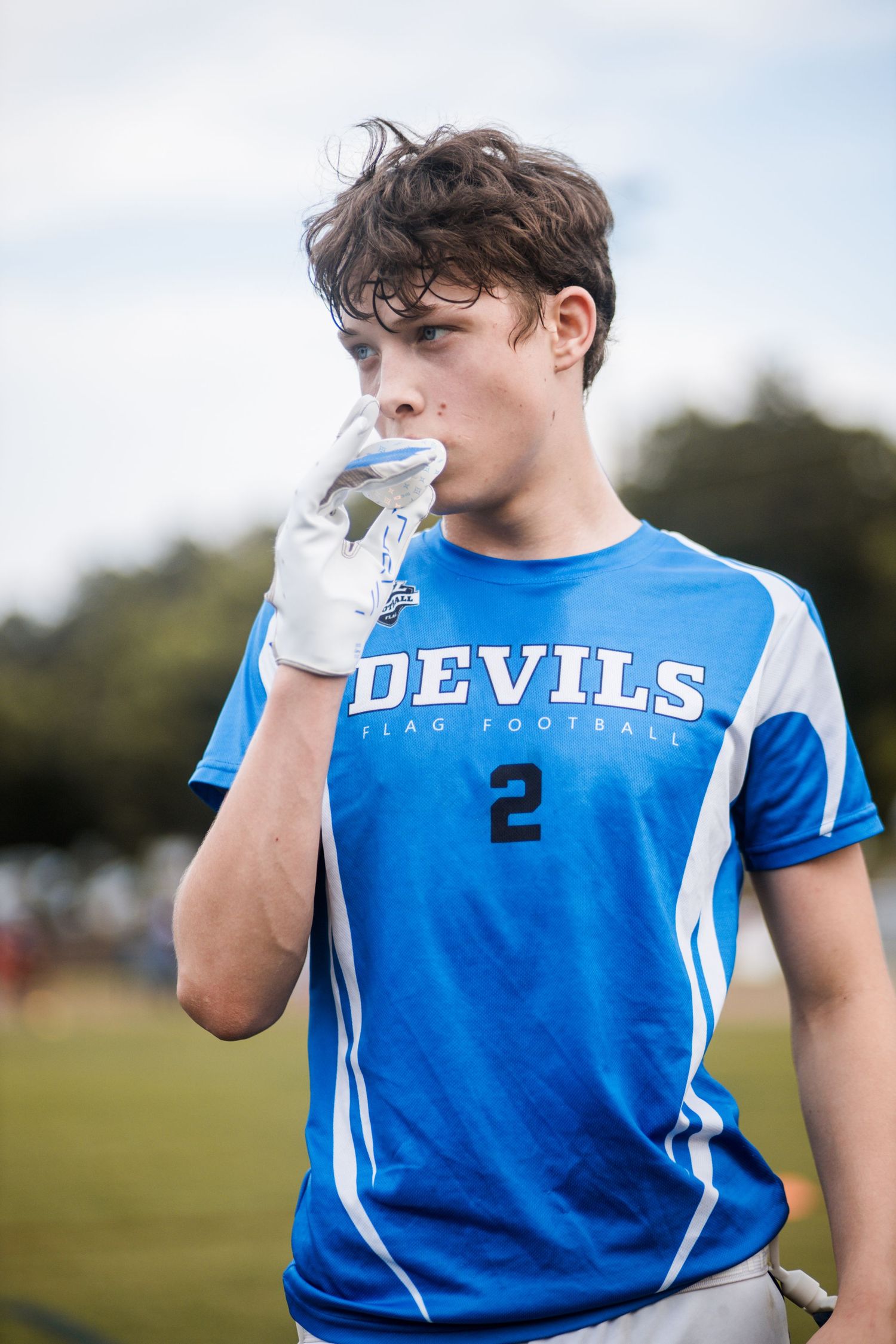 Teen boy in Devils #2 blue flag football uniform with gloved hand near mouth.