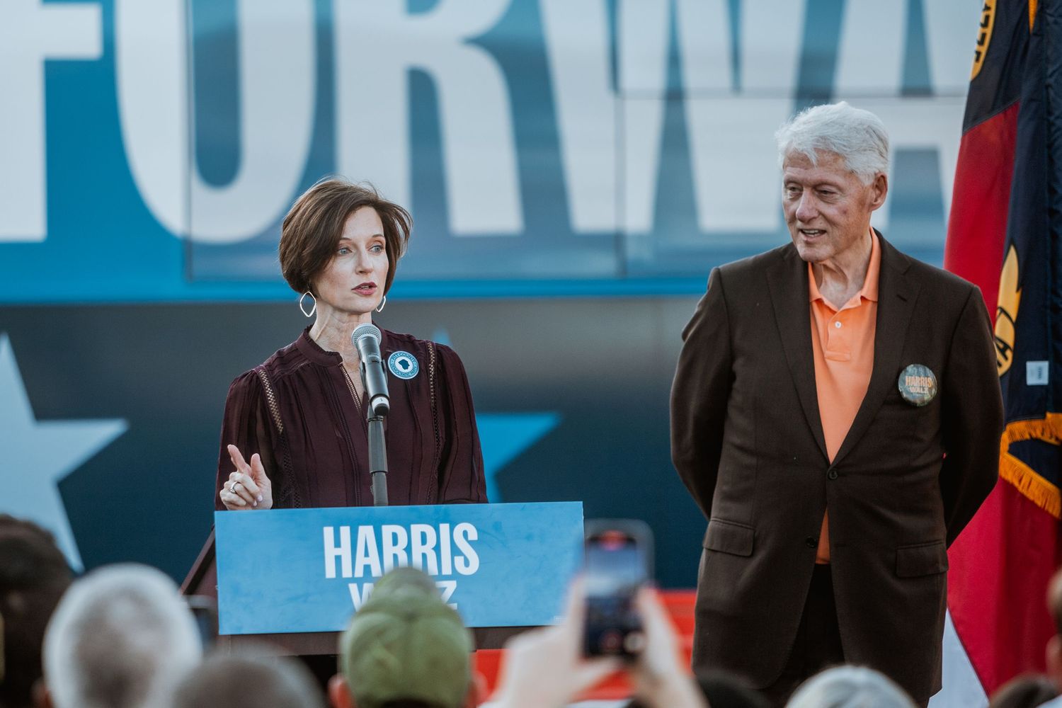 Two speakers at a Harris Walz campaign rally podium, with a blue Forward banner and North Carolina flag in background.