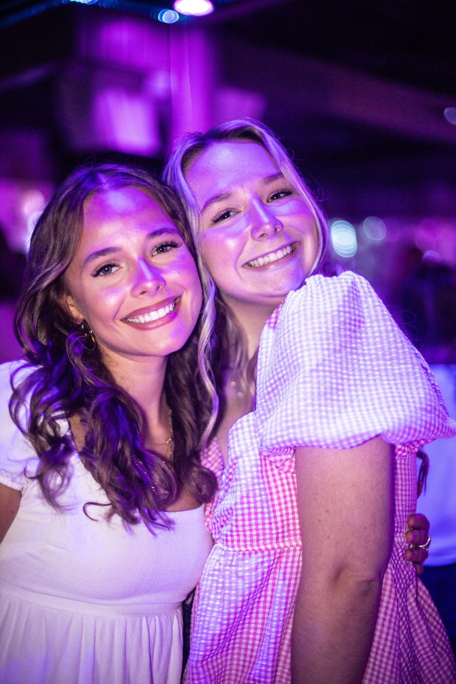 Two young women smile together under purple lighting at an indoor event.