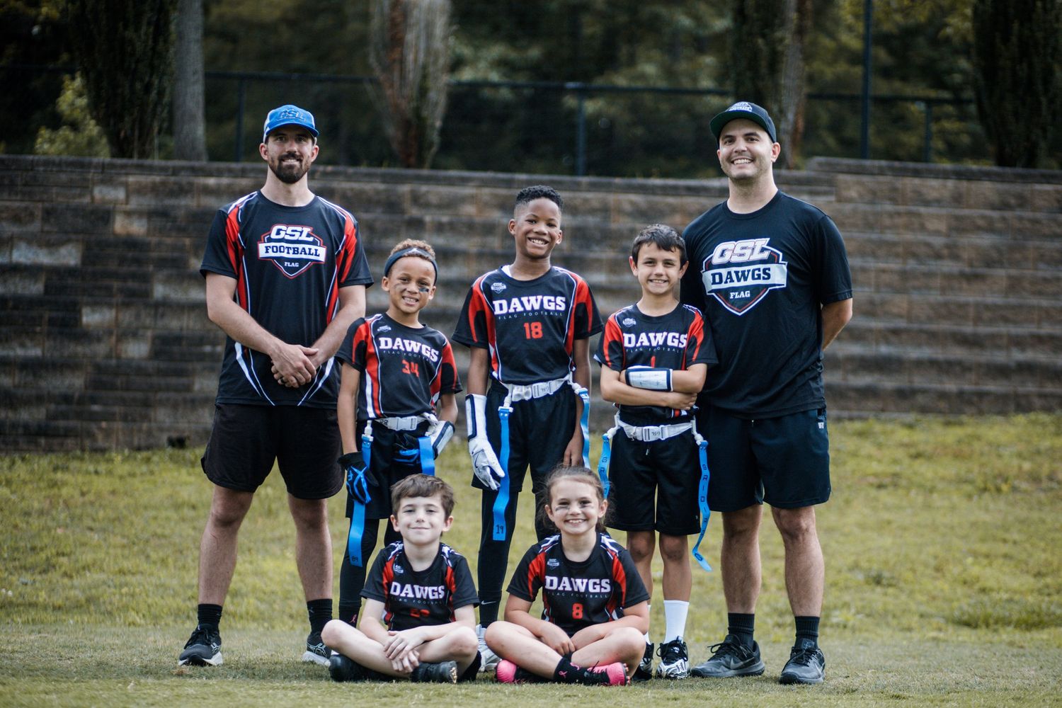 Small youth flag football team in black Dawgs uniforms posing with two coaches outdoors.