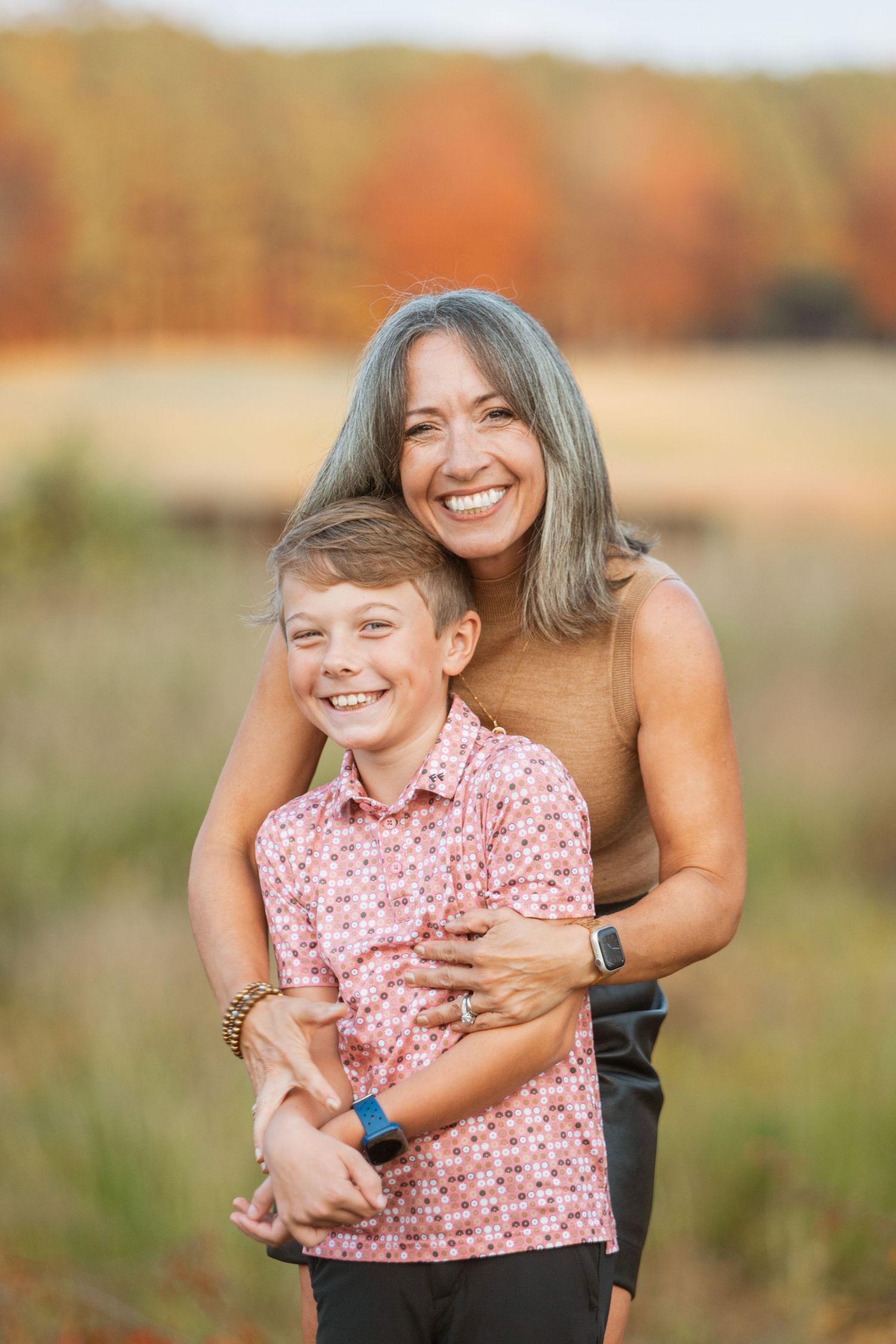 Laughing mother embraces young boy in pink floral shirt during golden autumn family portrait.