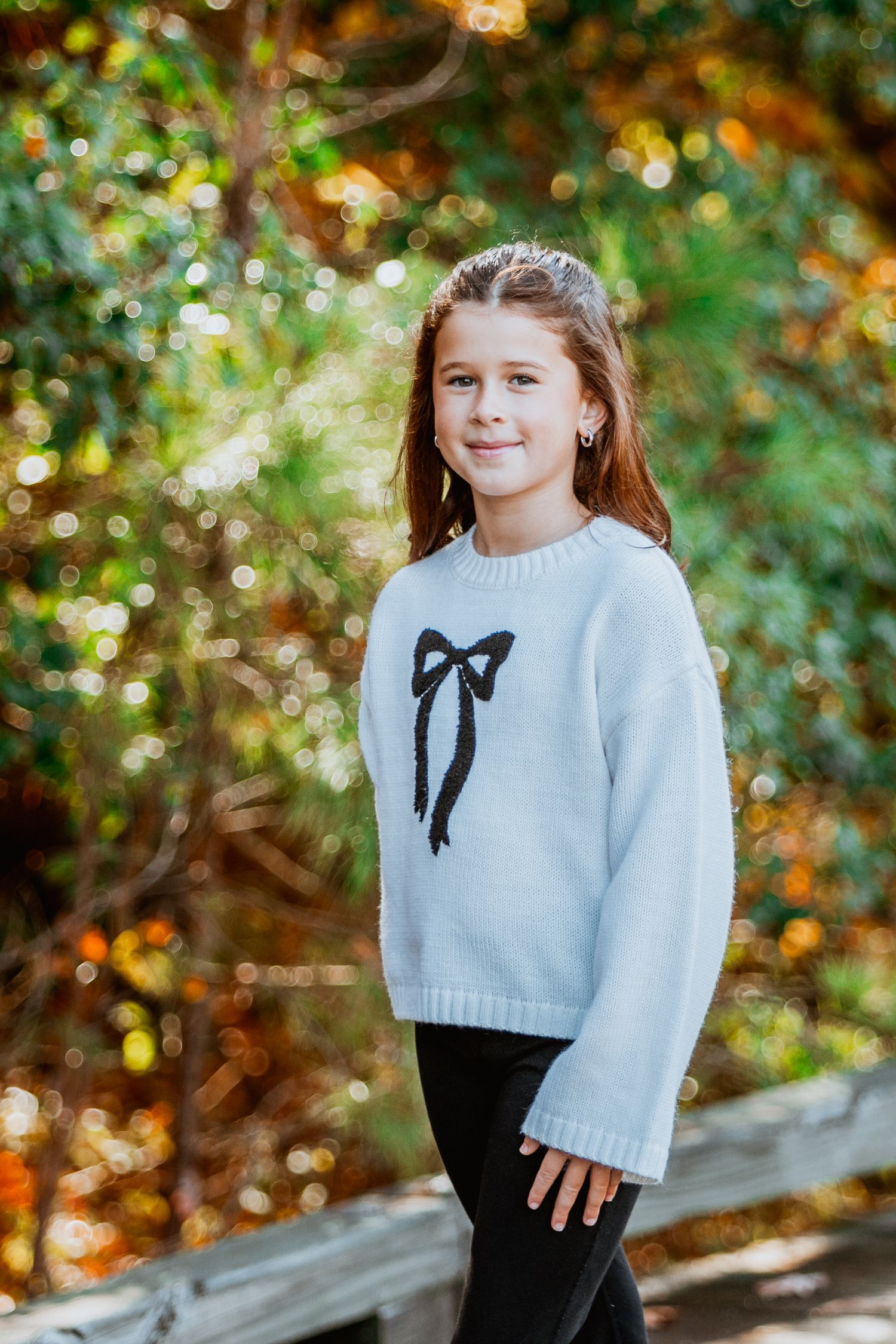 Young girl in white bow sweater and black pants smiling during autumn outdoor portrait.