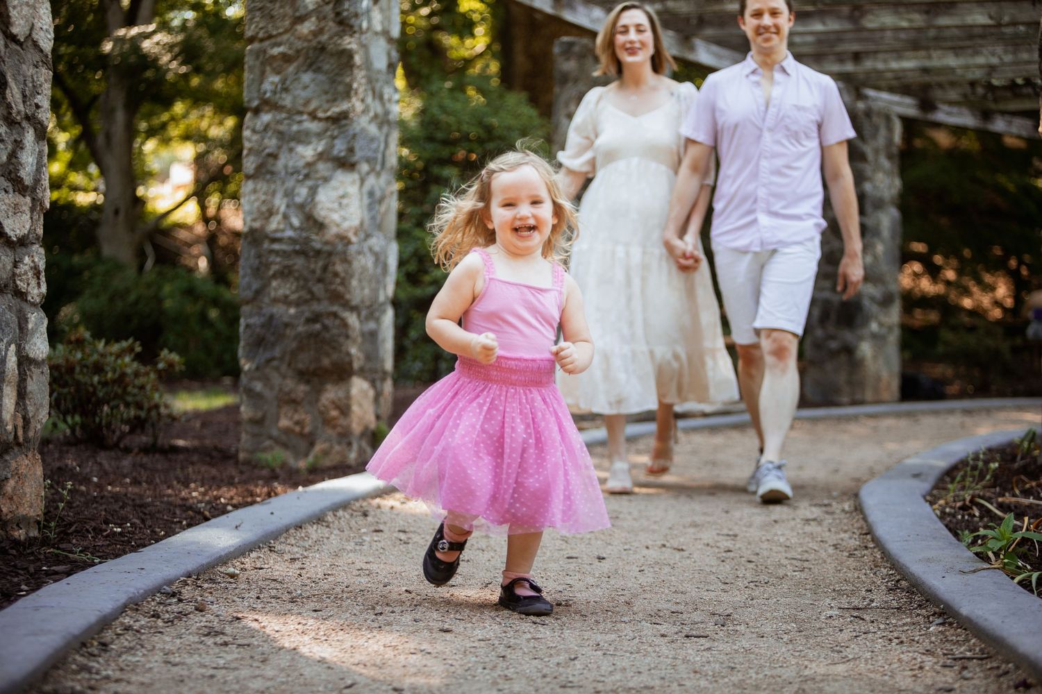 Smiling toddler in pink tutu runs along garden path while parents stroll behind under stone pergola.