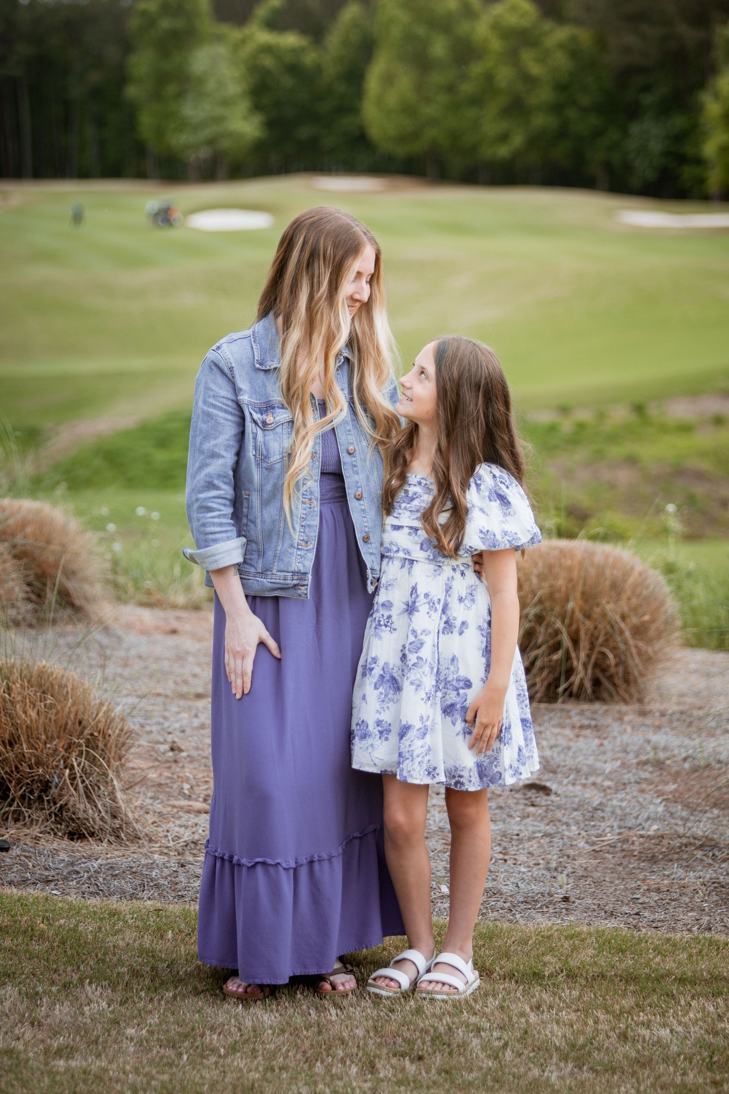Mother and daughter in purple and floral dresses smiling at each other on a golf course.