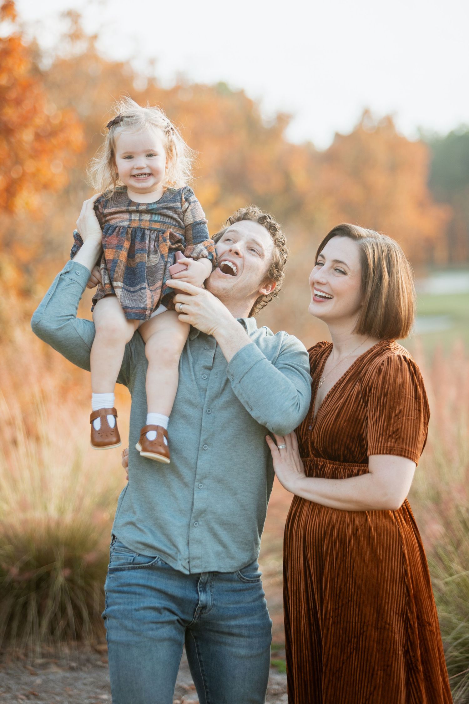 Laughing family of three with dad lifting toddler girl enjoys playful moment outdoors among warm autumn grasses.