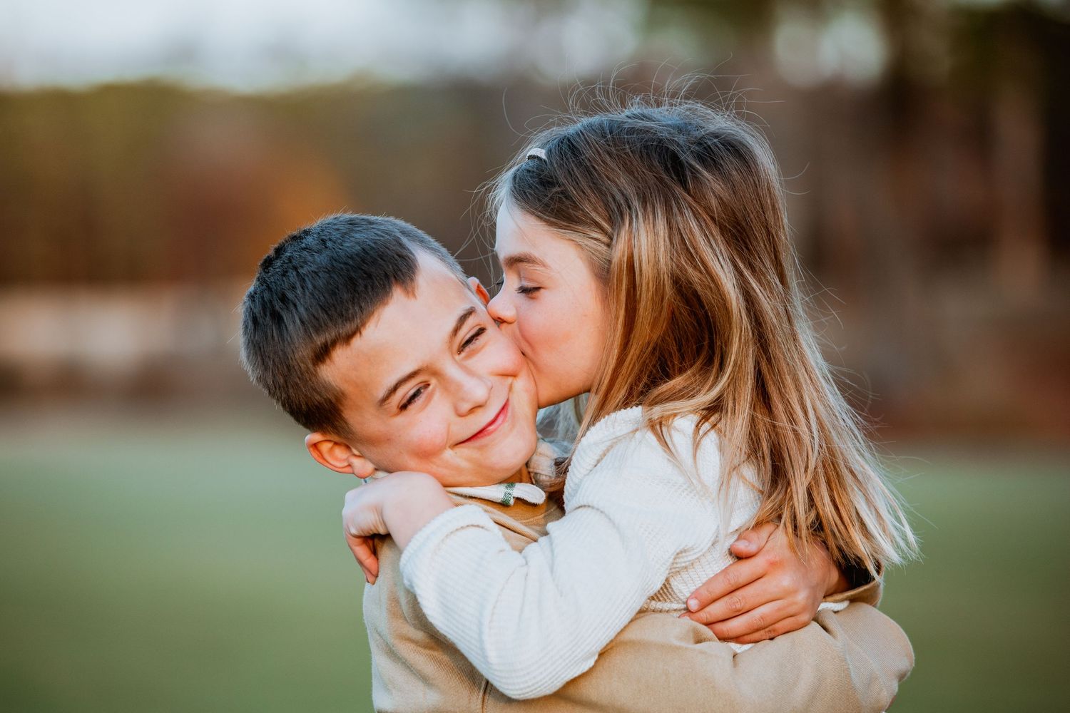 Girl kissing boy on the cheek while embracing outdoors in warm light.