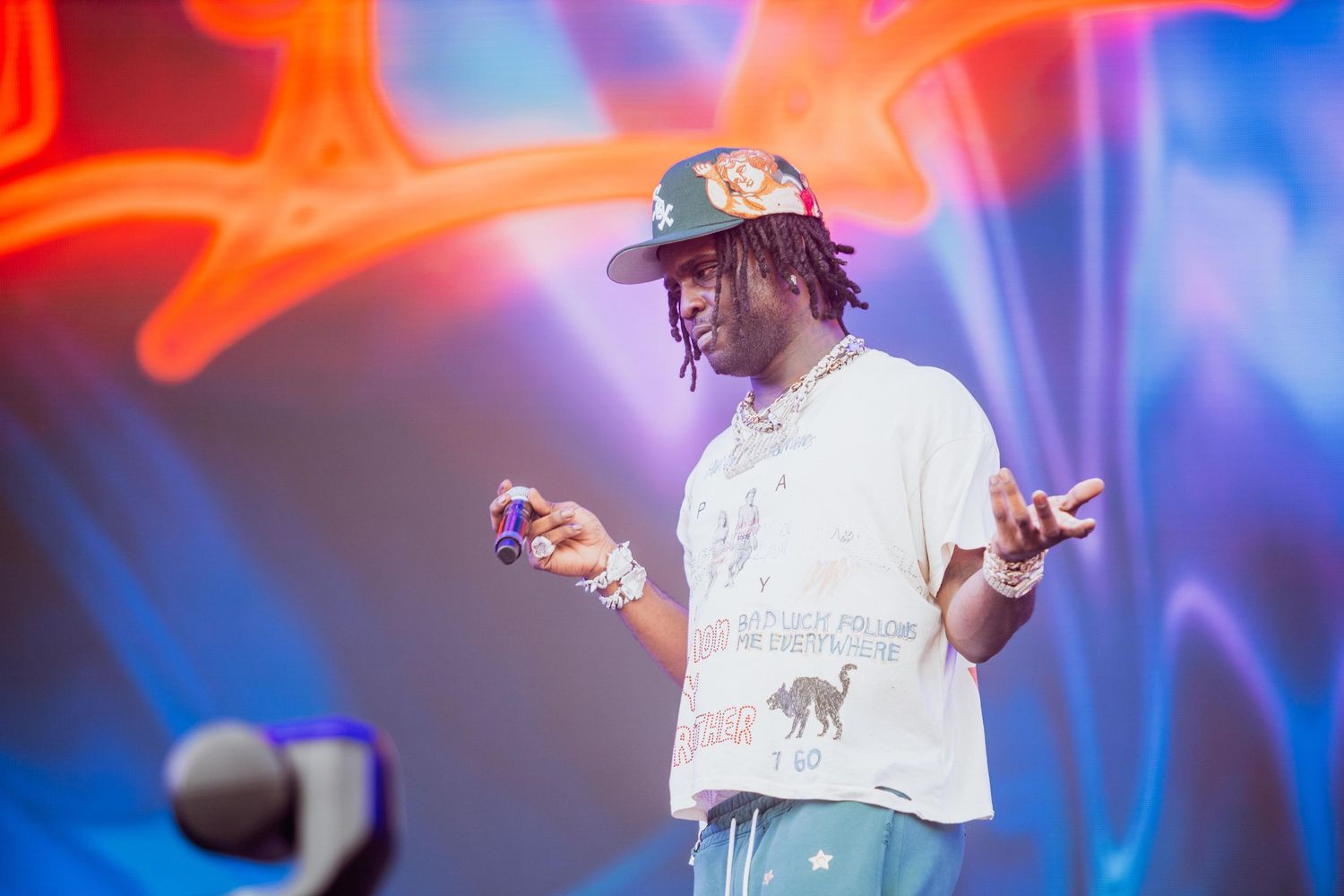 Young rapper on stage in a white graphic tee and cap gesturing with hands at a colorful outdoor concert.
