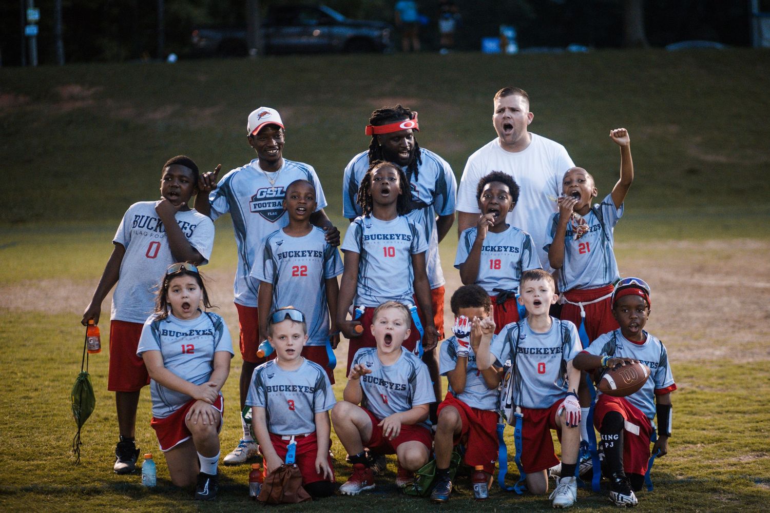 Youth flag football team in blue uniforms celebrating with coaches on a grass field at night.