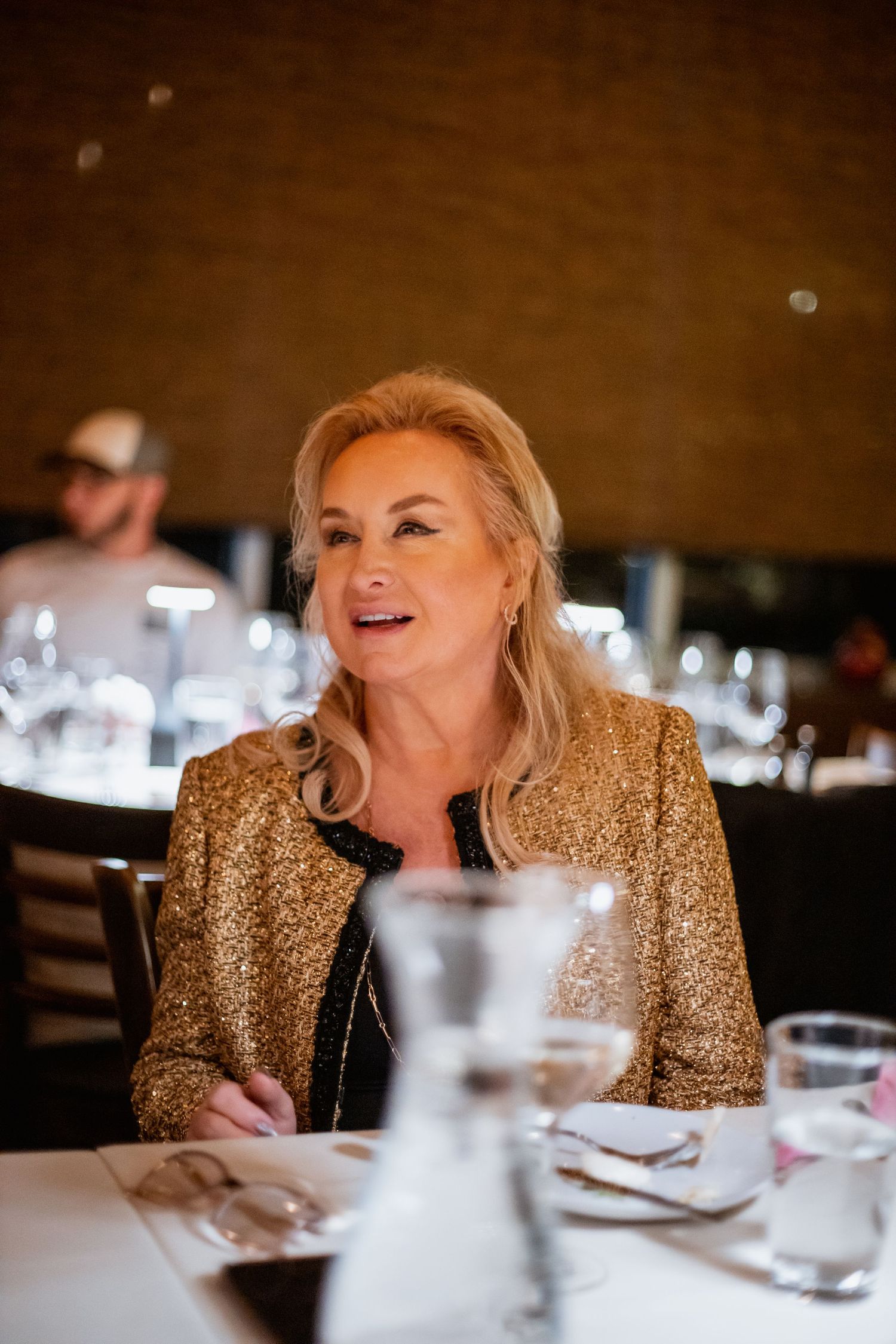 Elegant blonde woman in gold sequin jacket dining at upscale restaurant with wine glasses on white tablecloth.