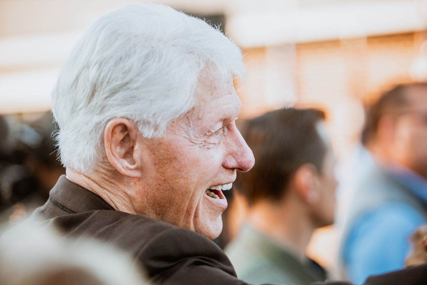 Elderly white-haired man smiles and laughs in profile at political event.