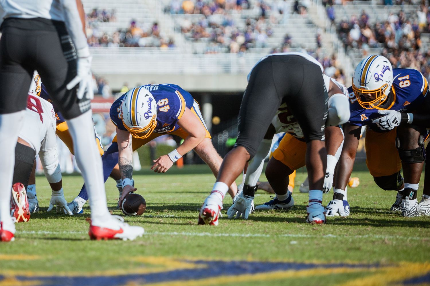 ECU Pirates offensive linemen prepare for snap in blue and gold uniforms during college football game.