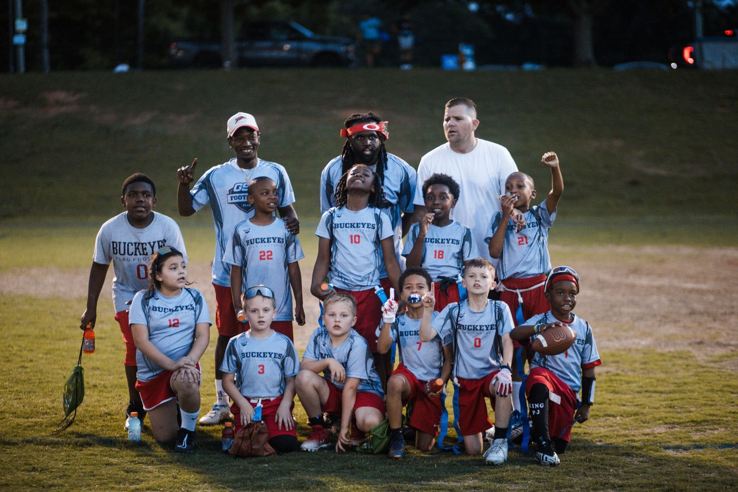 Youth flag football team in light blue uniforms posing with coaches on a grass field at dusk.
