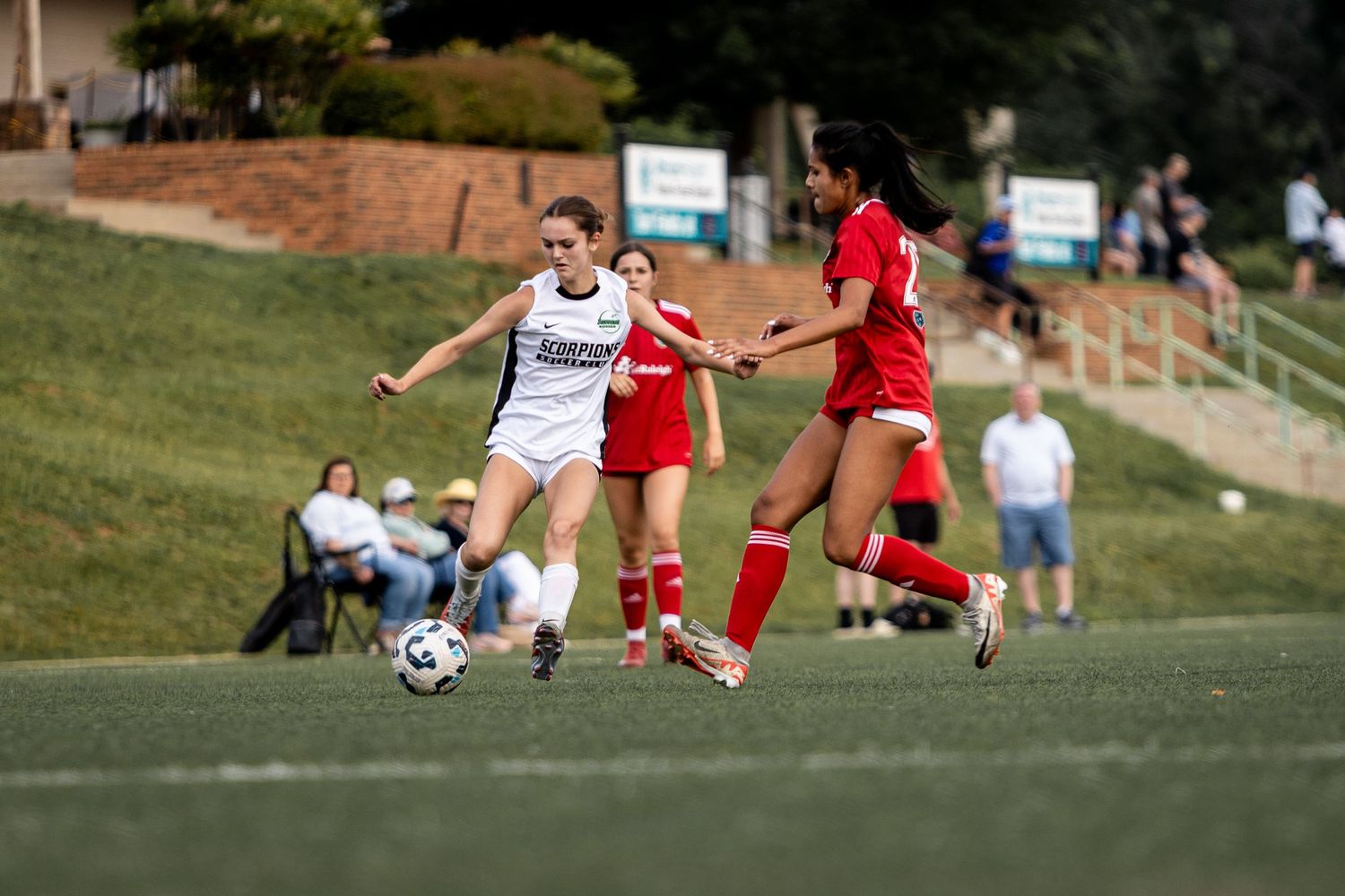 Female soccer player in white dribbles past defenders in red during an outdoor soccer game.