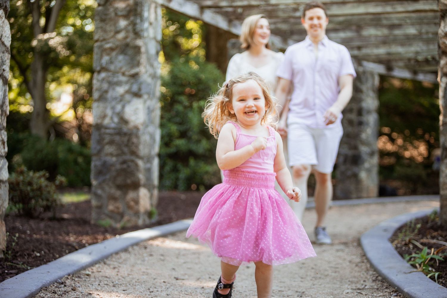 Happy little girl in pink dress running toward camera with parents walking behind in a garden path.