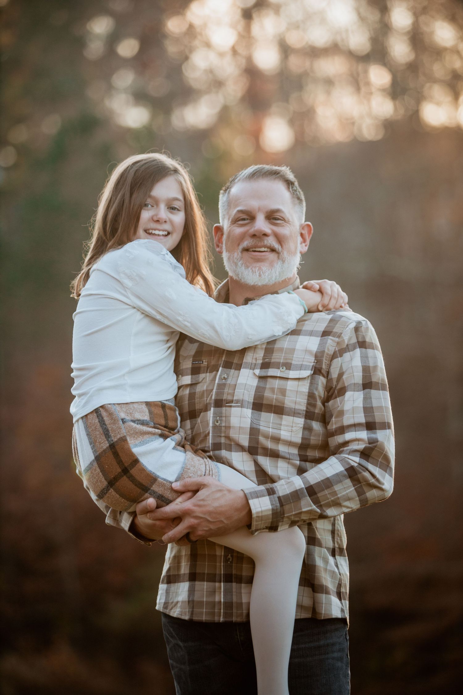 Smiling bearded father holding his daughter in his arms outdoors in warm golden light.