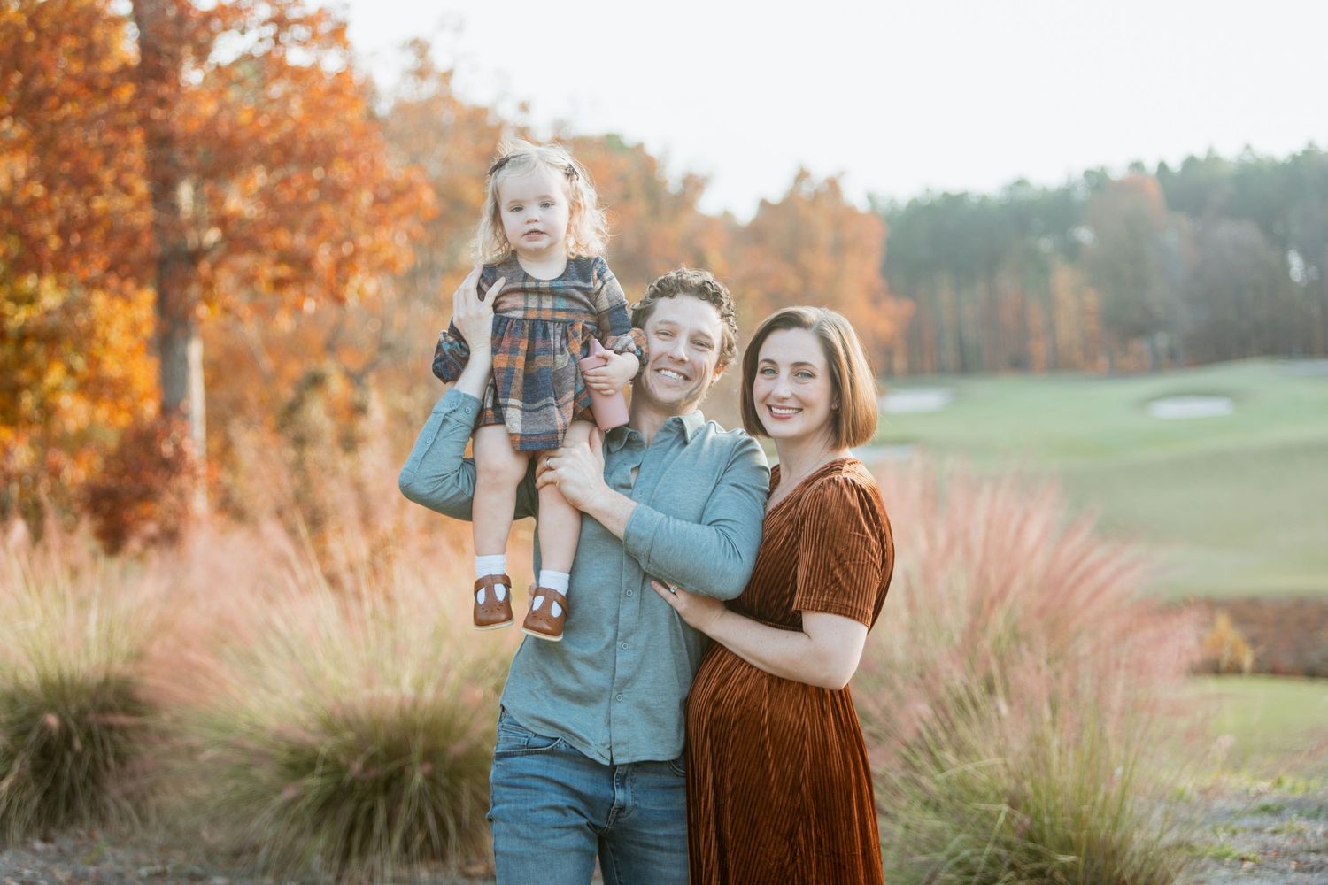Joyful family poses with dad holding toddler on shoulders and mom beside them among vibrant fall foliage on golf course.