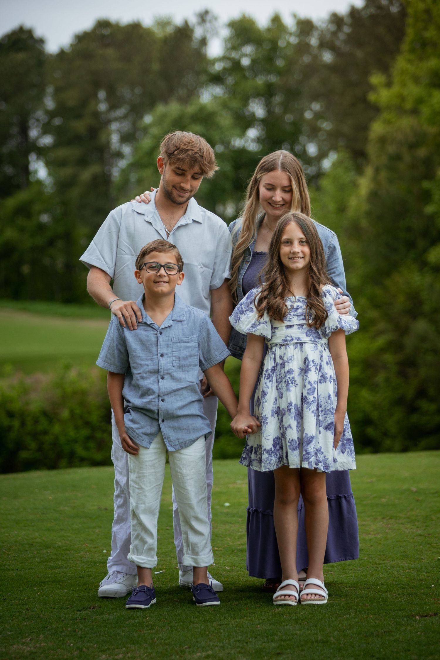 Family of four posing together on green grass with tall trees in the background.