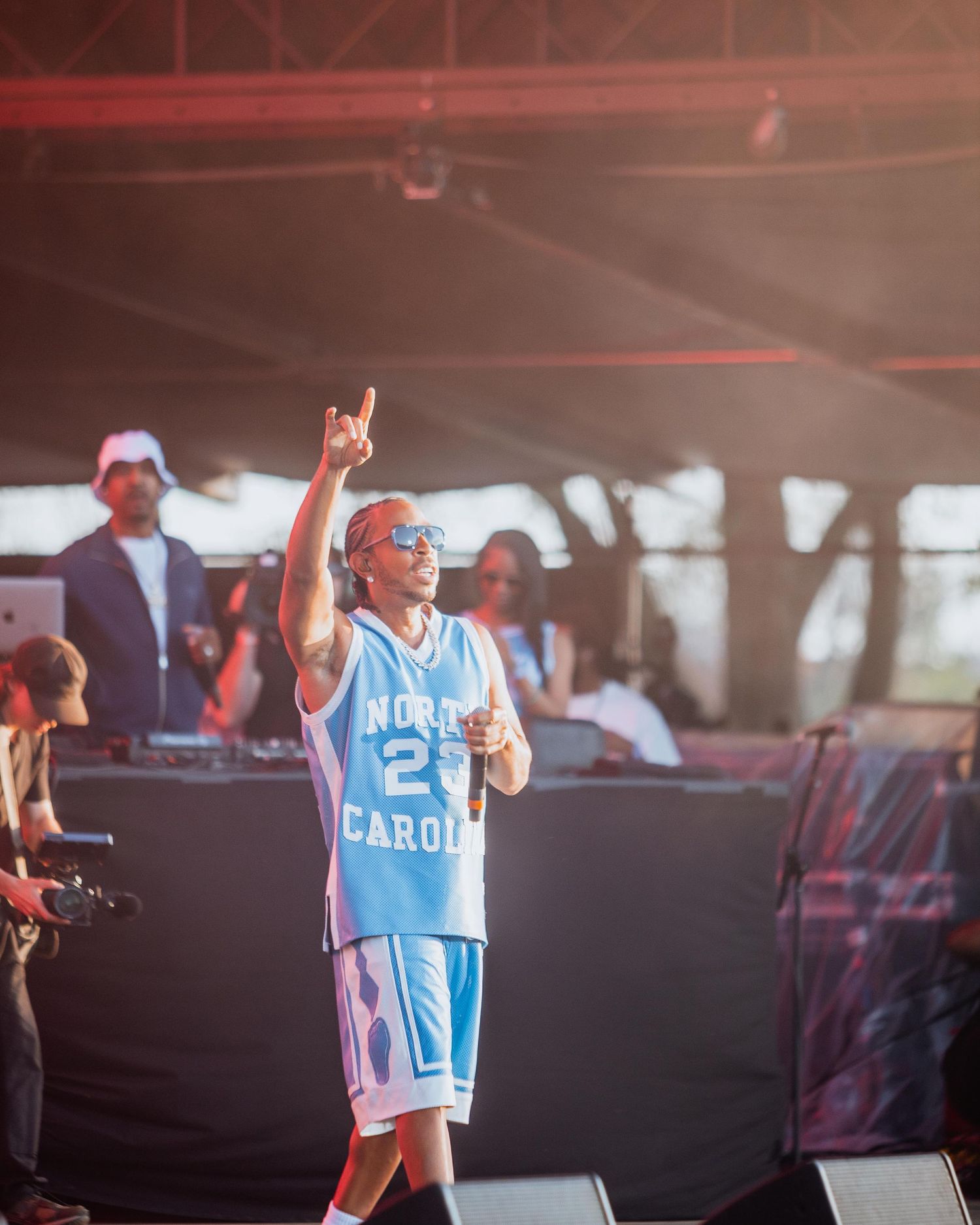 Artist in a light blue North Carolina basketball jersey pointing upward while performing at an outdoor festival.