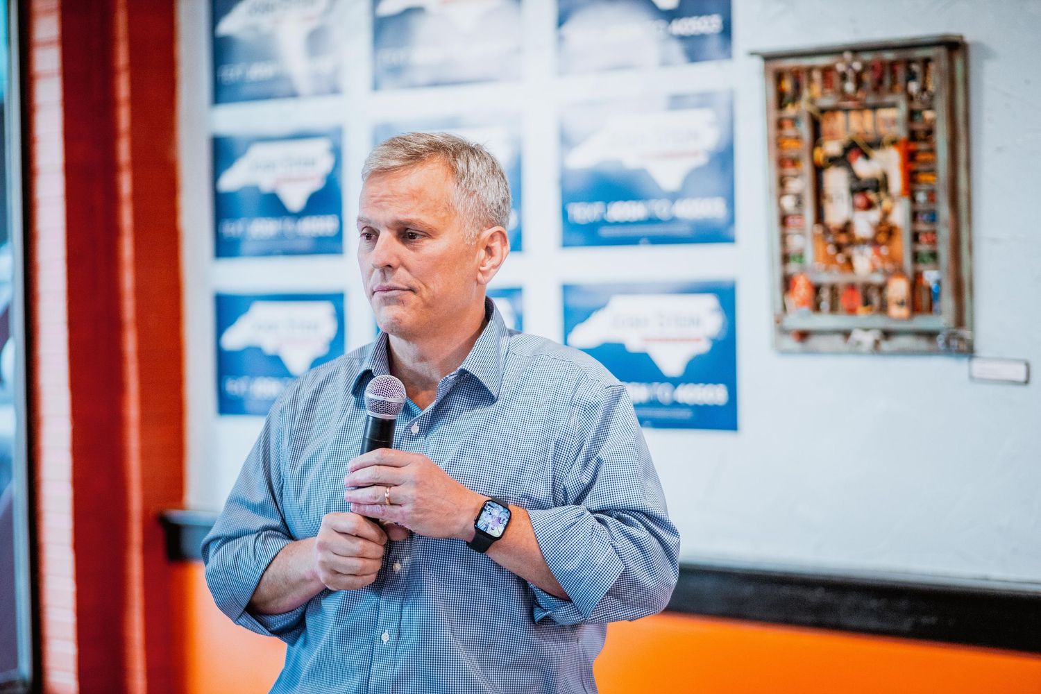 Man holding microphone speaking at indoor North Carolina political campaign rally.
