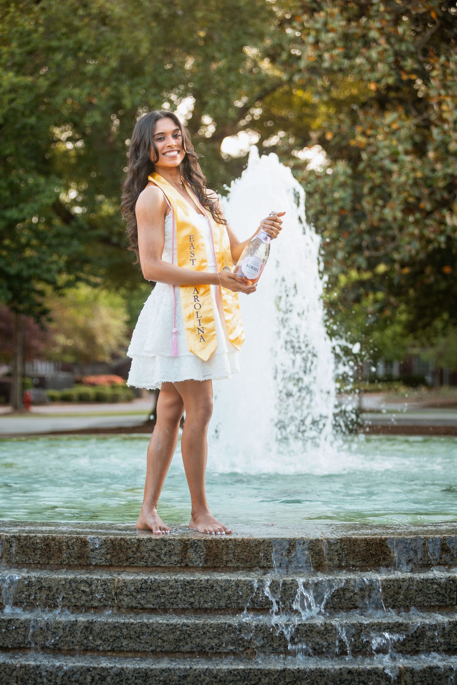 Joyful graduate stands barefoot on fountain steps holding champagne bottle with East Carolina sash.