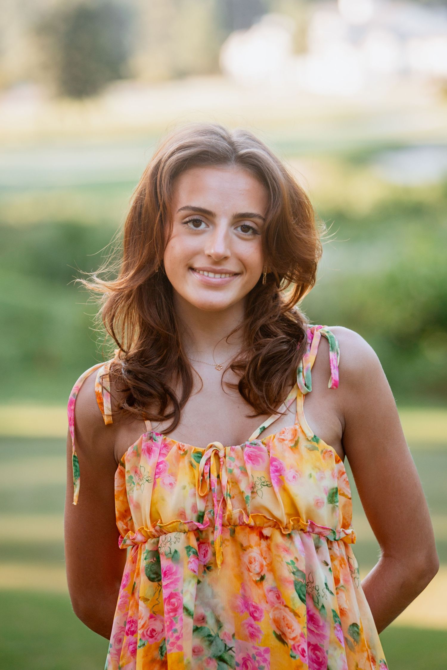 Smiling teenage girl with long brown hair wearing floral sundress in outdoor setting.