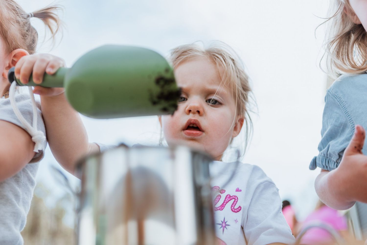 Curious toddler girl holding a green bottle at an outdoor play area on a bright day.