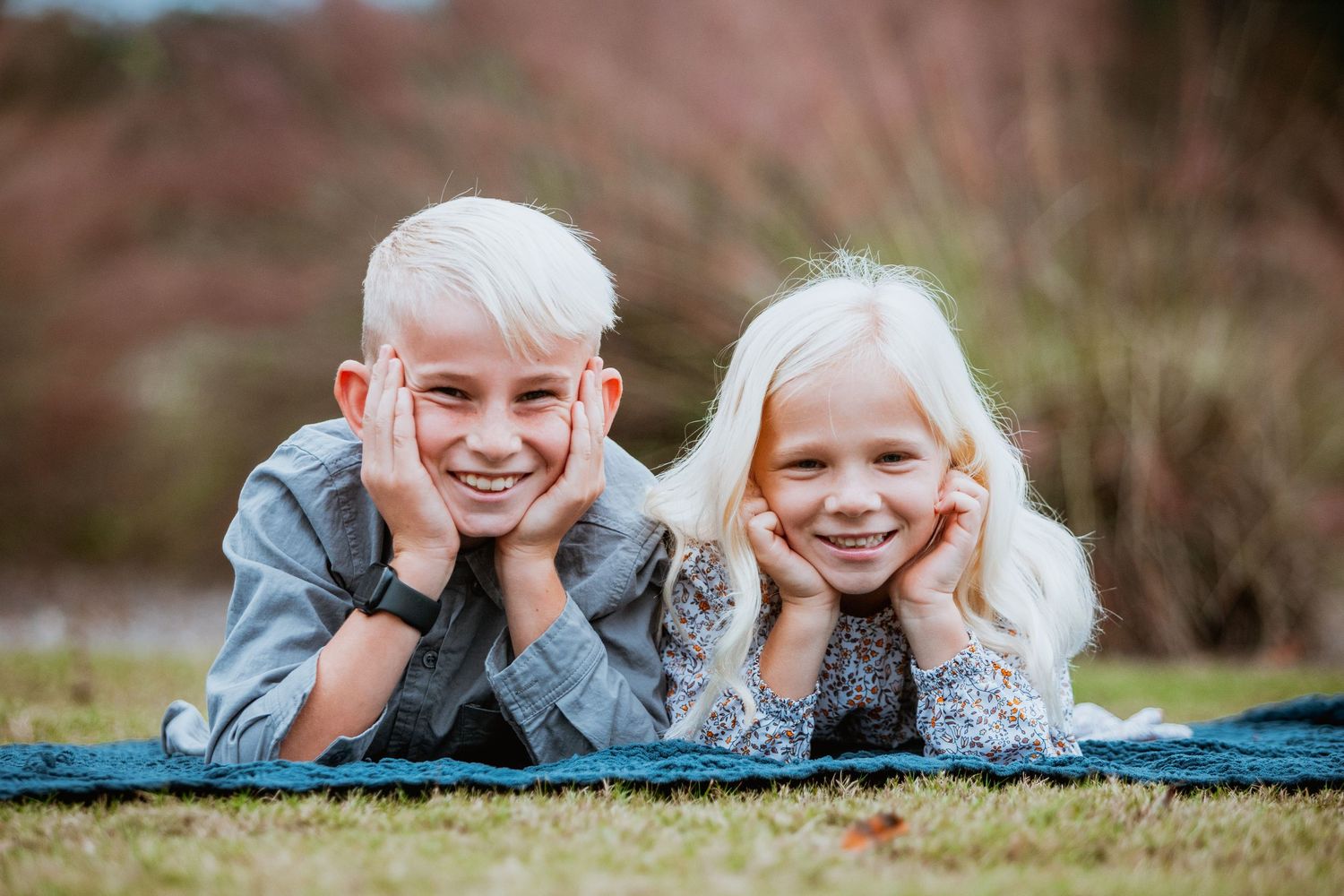 Two blonde siblings lie on grass smiling with chins resting on hands during outdoor photo session.