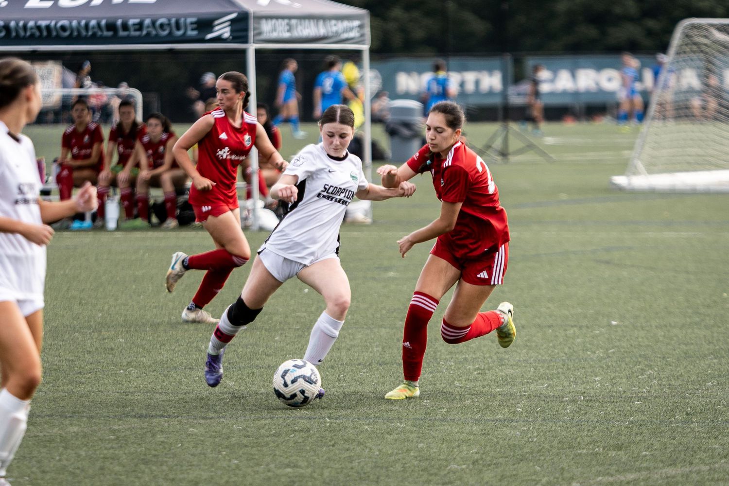 Female soccer player in white Scorpions kit dribbles past red-uniformed defender during National League match.