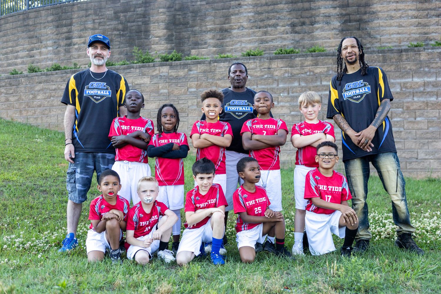 Youth flag football team in red jerseys poses with two coaches for team photo against stone wall.
