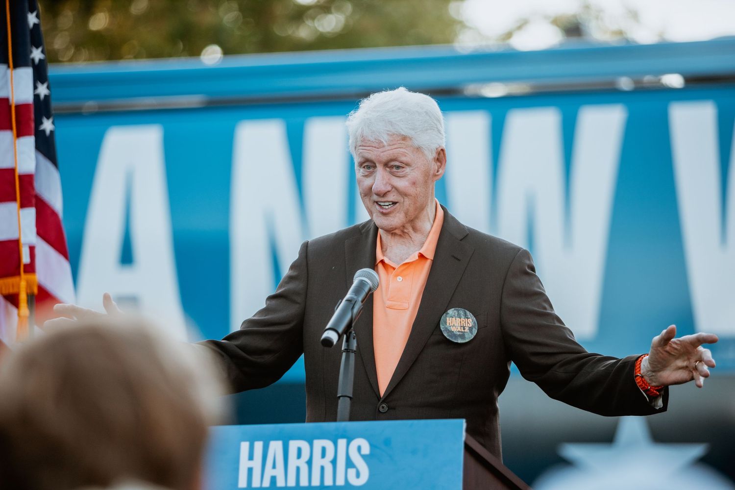 Older man in orange shirt gestures while speaking at Harris-Walz campaign rally with American flag nearby.