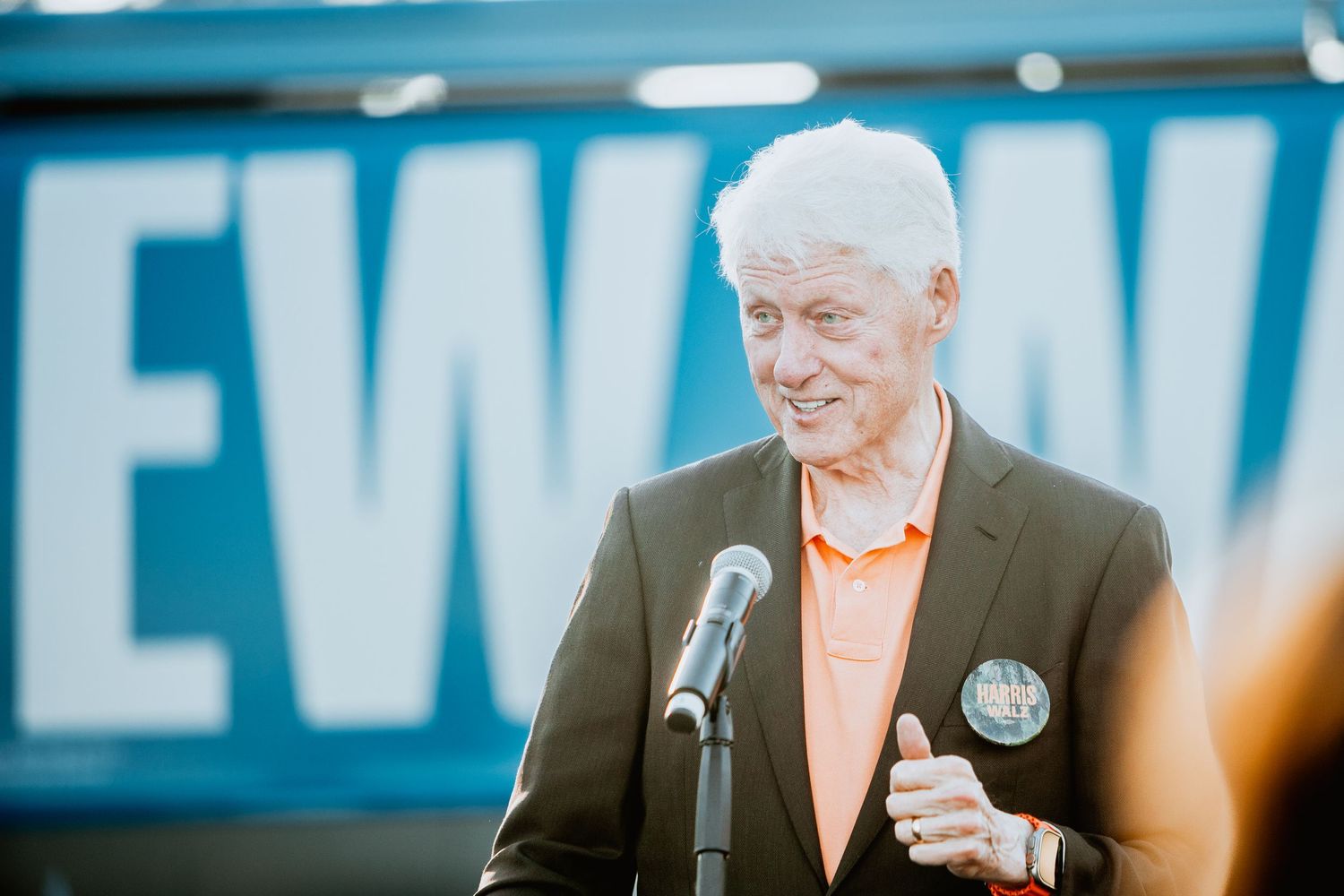 Senior man with white hair smiling at podium in dark blazer and orange shirt at outdoor rally event.