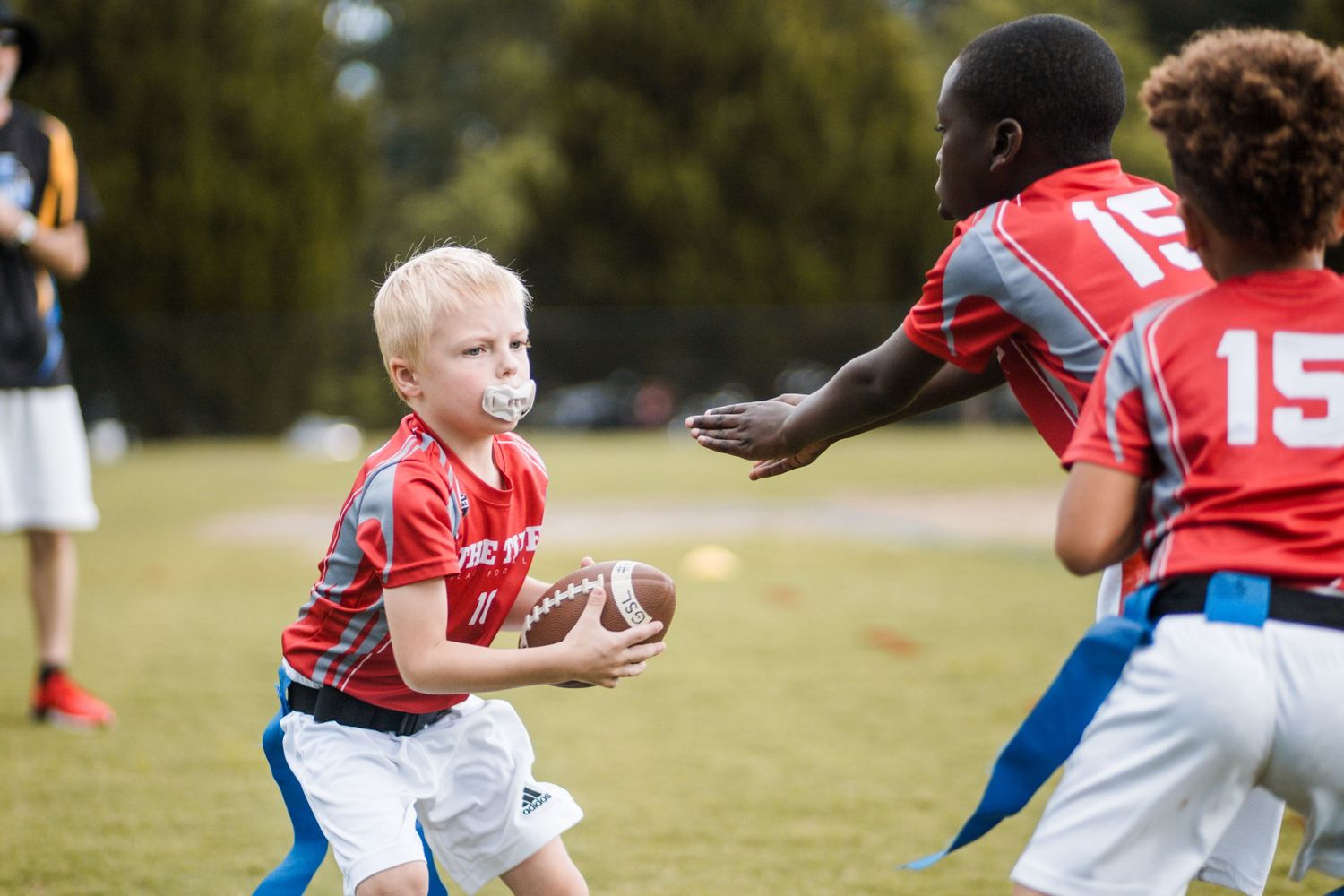 Young blonde boy holds football while defender reaches for flag in youth flag football game.