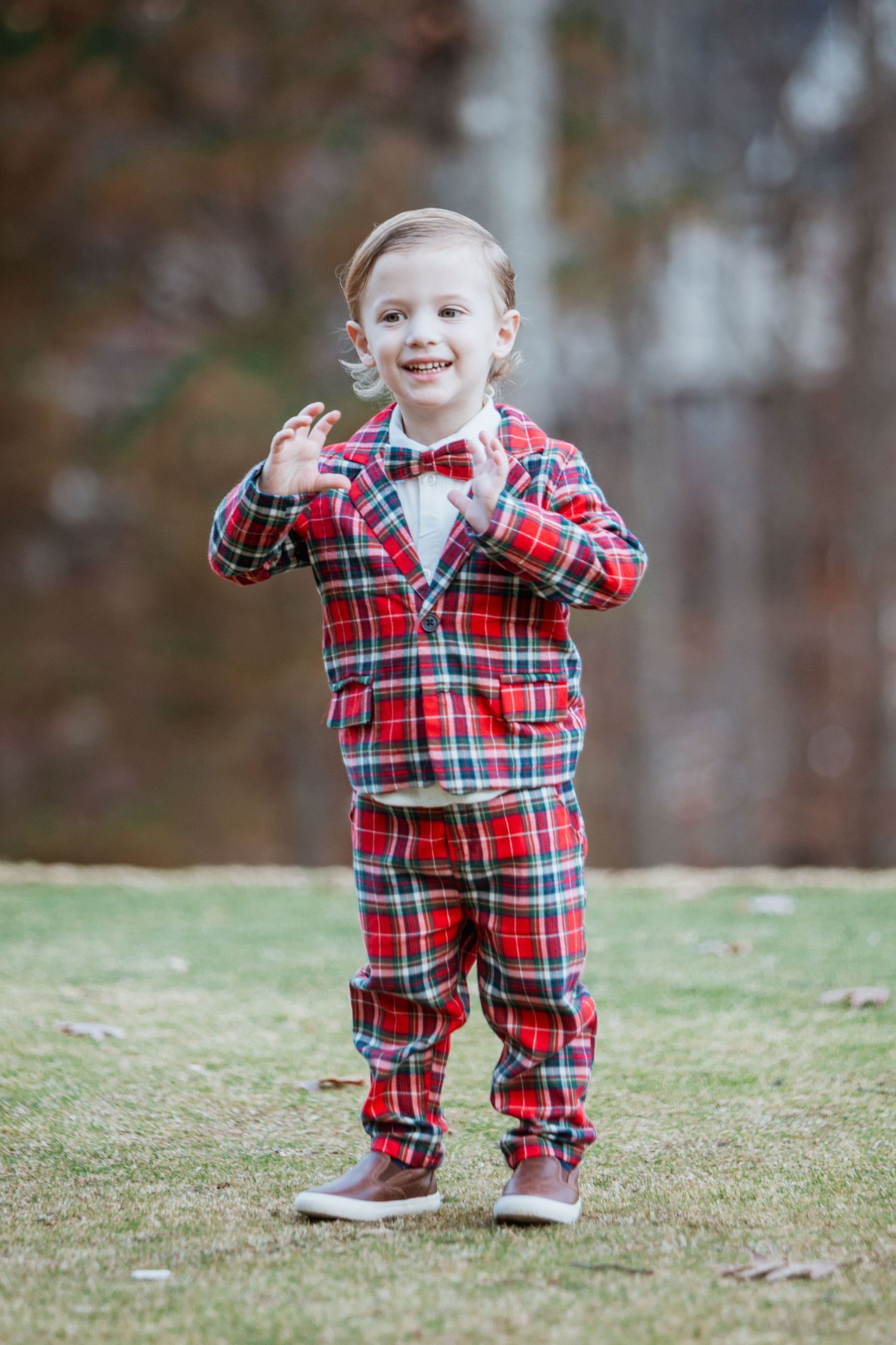 Smiling toddler boy in red plaid suit with bow tie clapping hands outdoors.