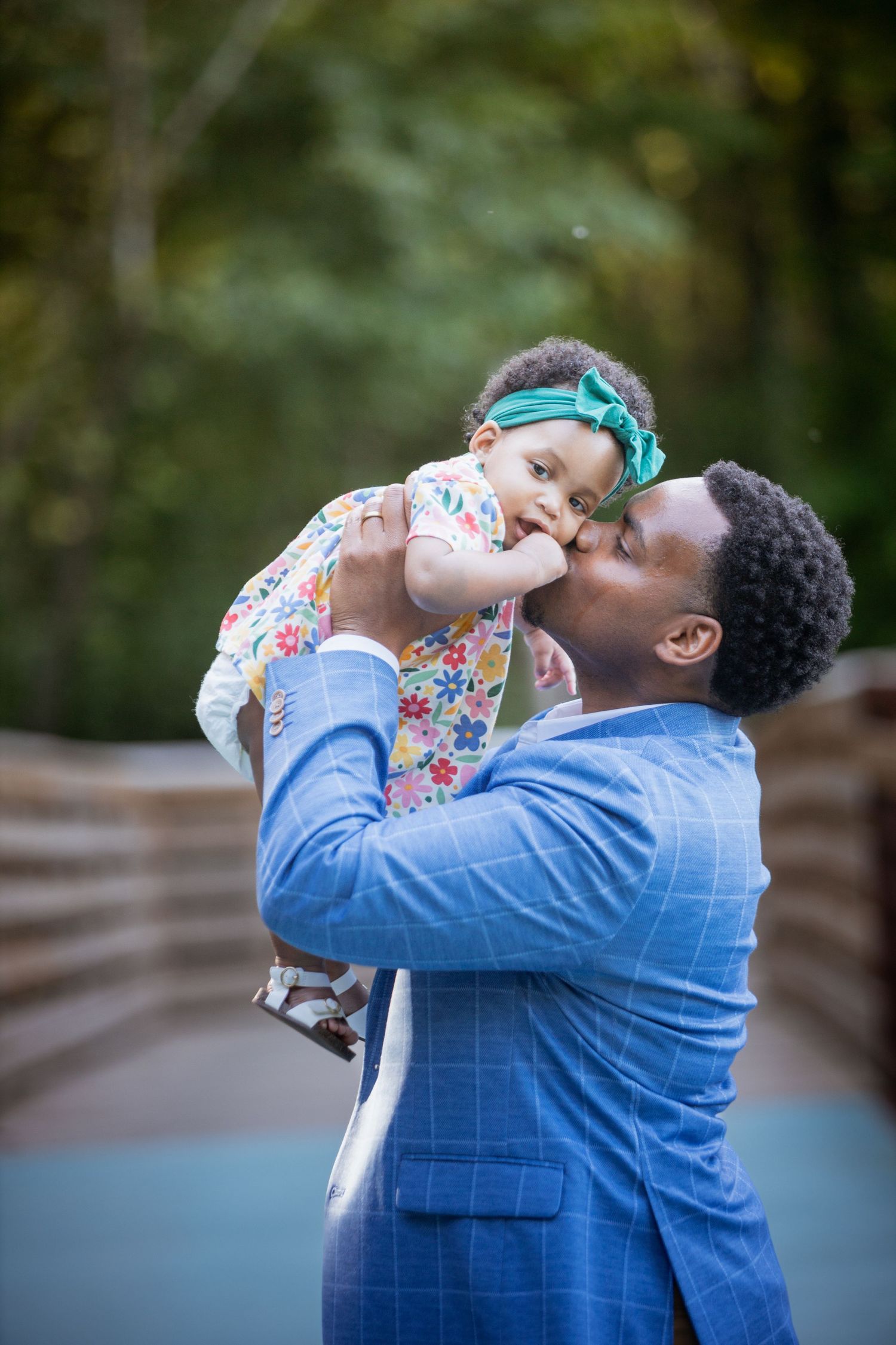 Father in blue blazer lovingly kissing his baby girl in floral dress while lifting her up outdoors.