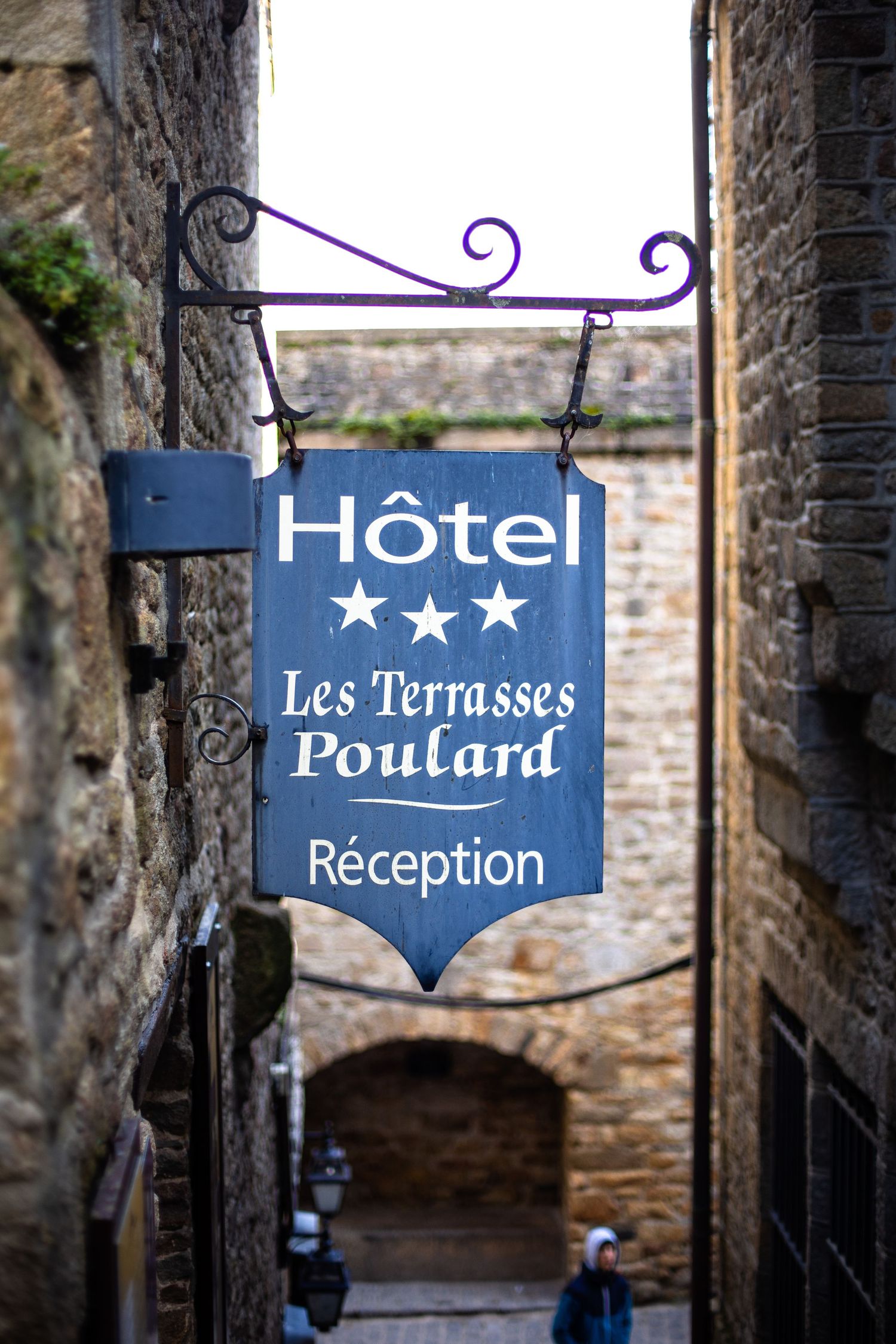 Blue hotel sign for Les Terrasses Poulard three-star hotel hanging in a narrow stone alley.