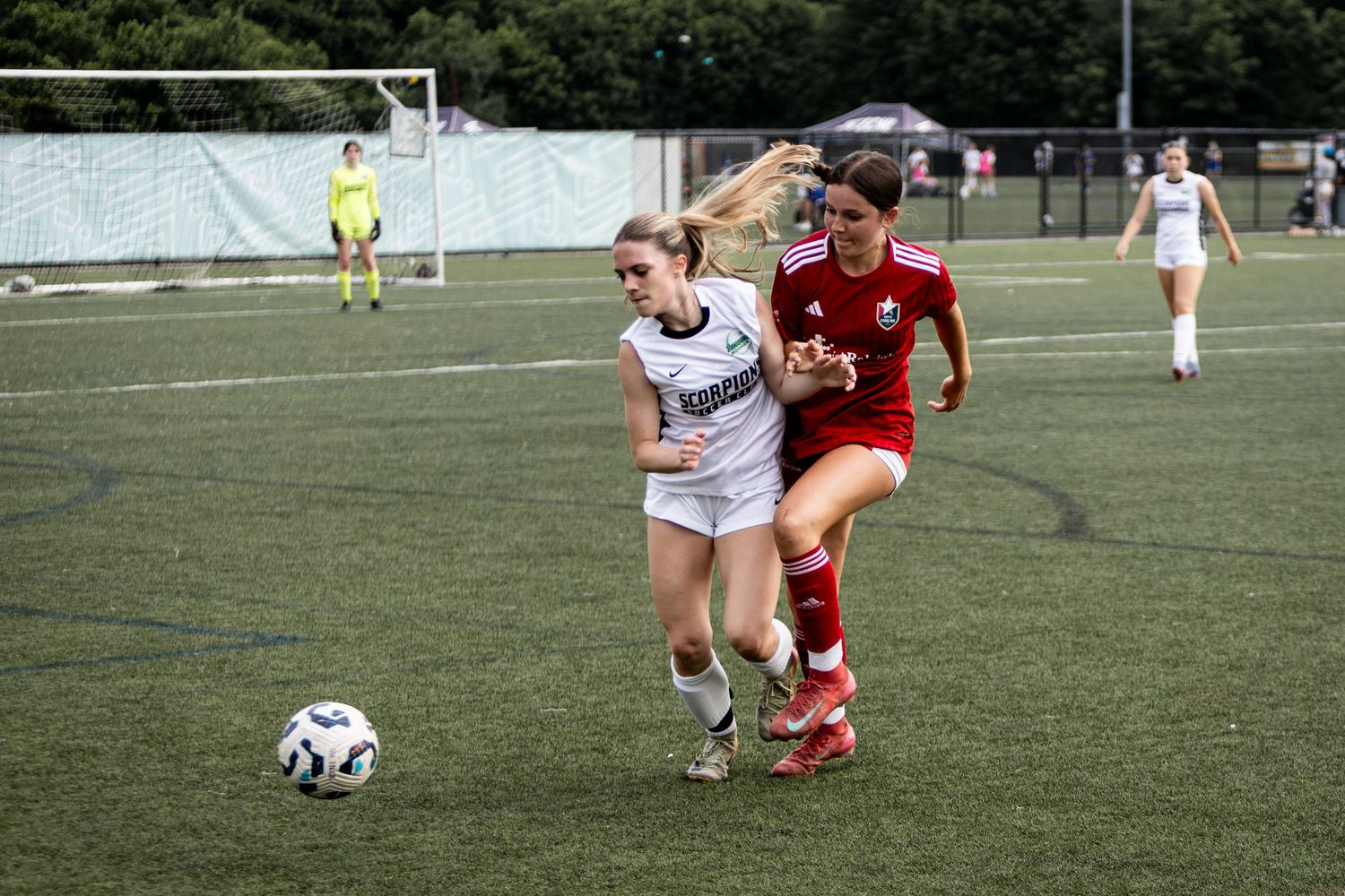 Two female soccer players compete for the ball during a match on a green turf field.