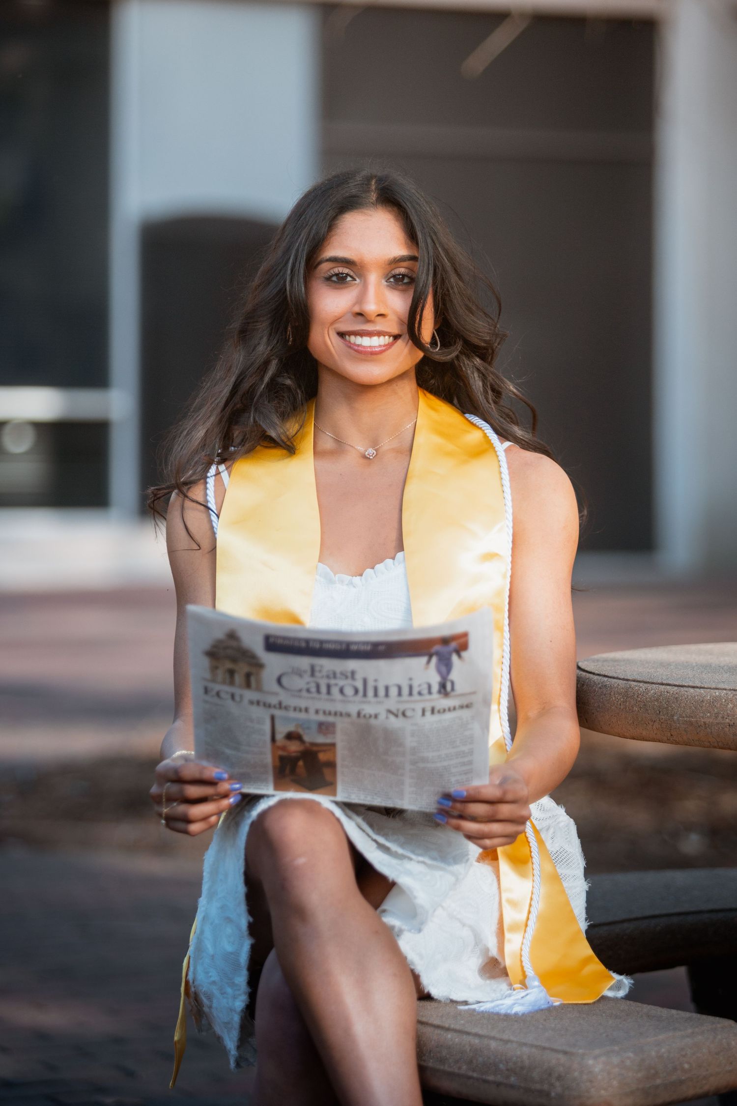 Smiling graduate in white dress holds East Carolinian newspaper while wearing gold graduation stole.