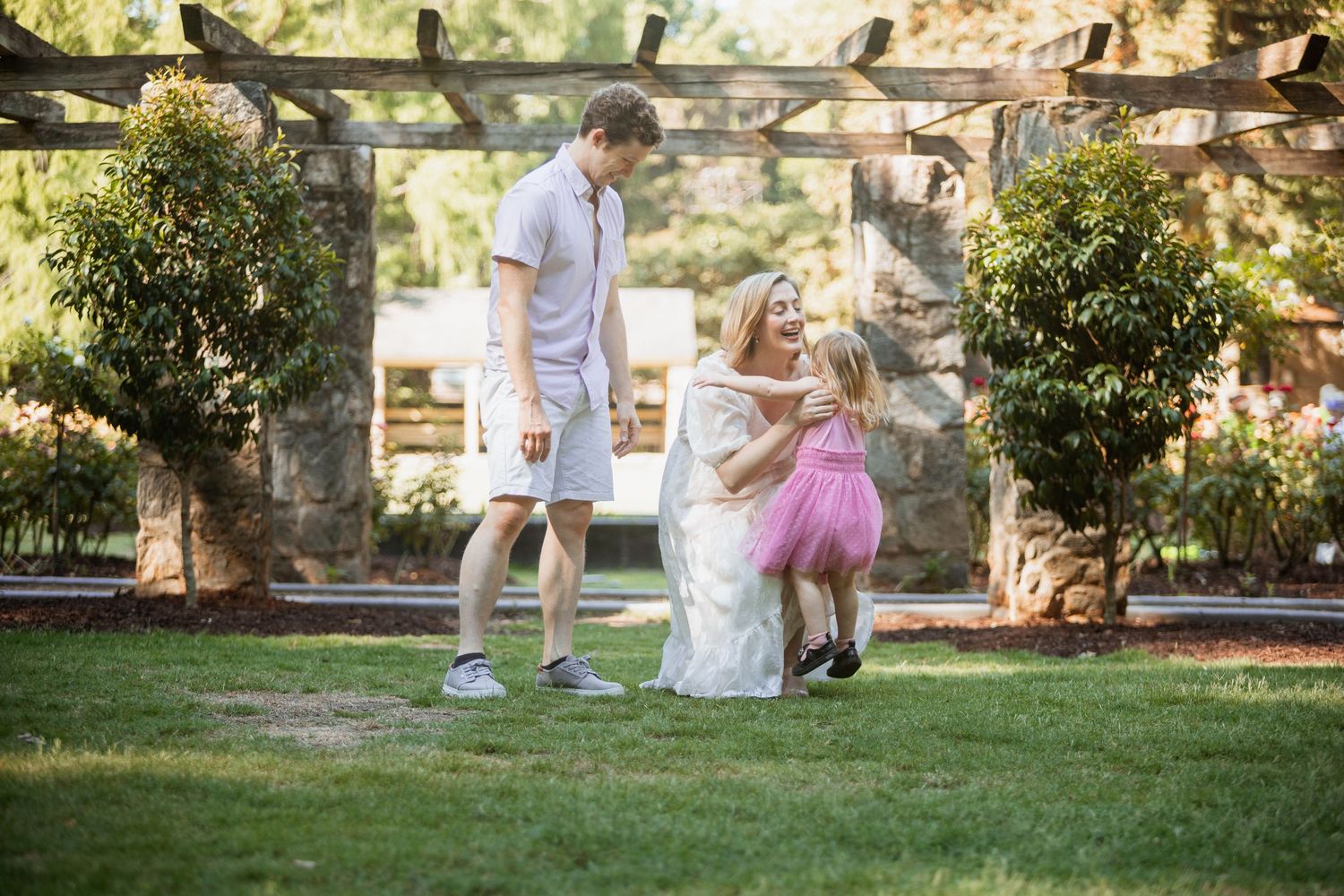 Family of three enjoying a sunny day in a lush garden, mom in white dress crouching to greet toddler in pink tutu.