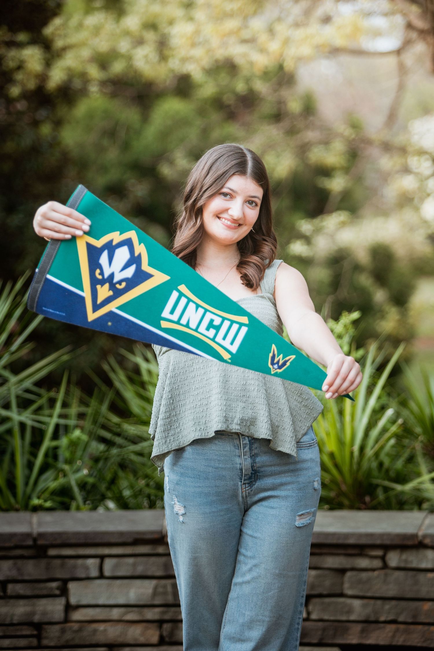 Smiling young woman holding a UNCW pennant flag outdoors in a garden setting.