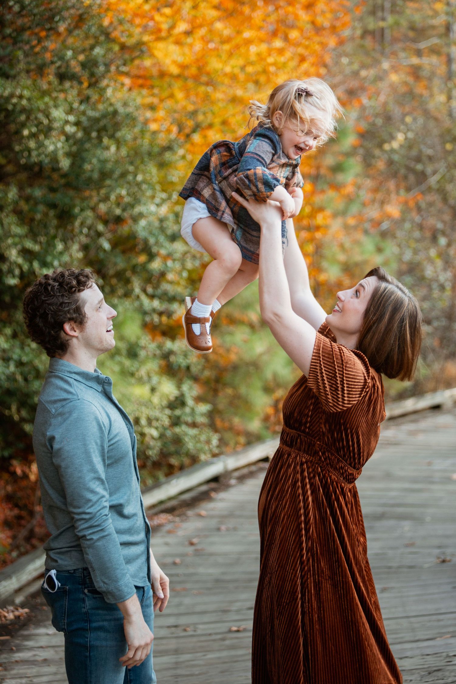Mom in rust velvet dress joyfully lifts laughing toddler overhead on wooden boardwalk surrounded by autumn foliage.