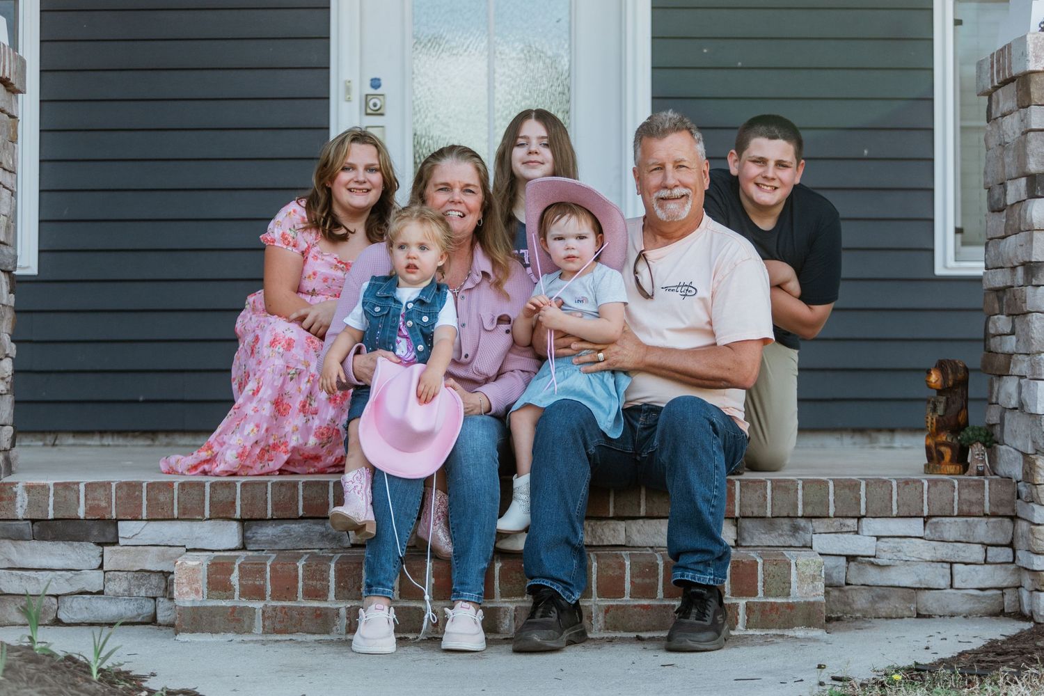 A smiling family of six poses together on brick front porch steps of a gray house.