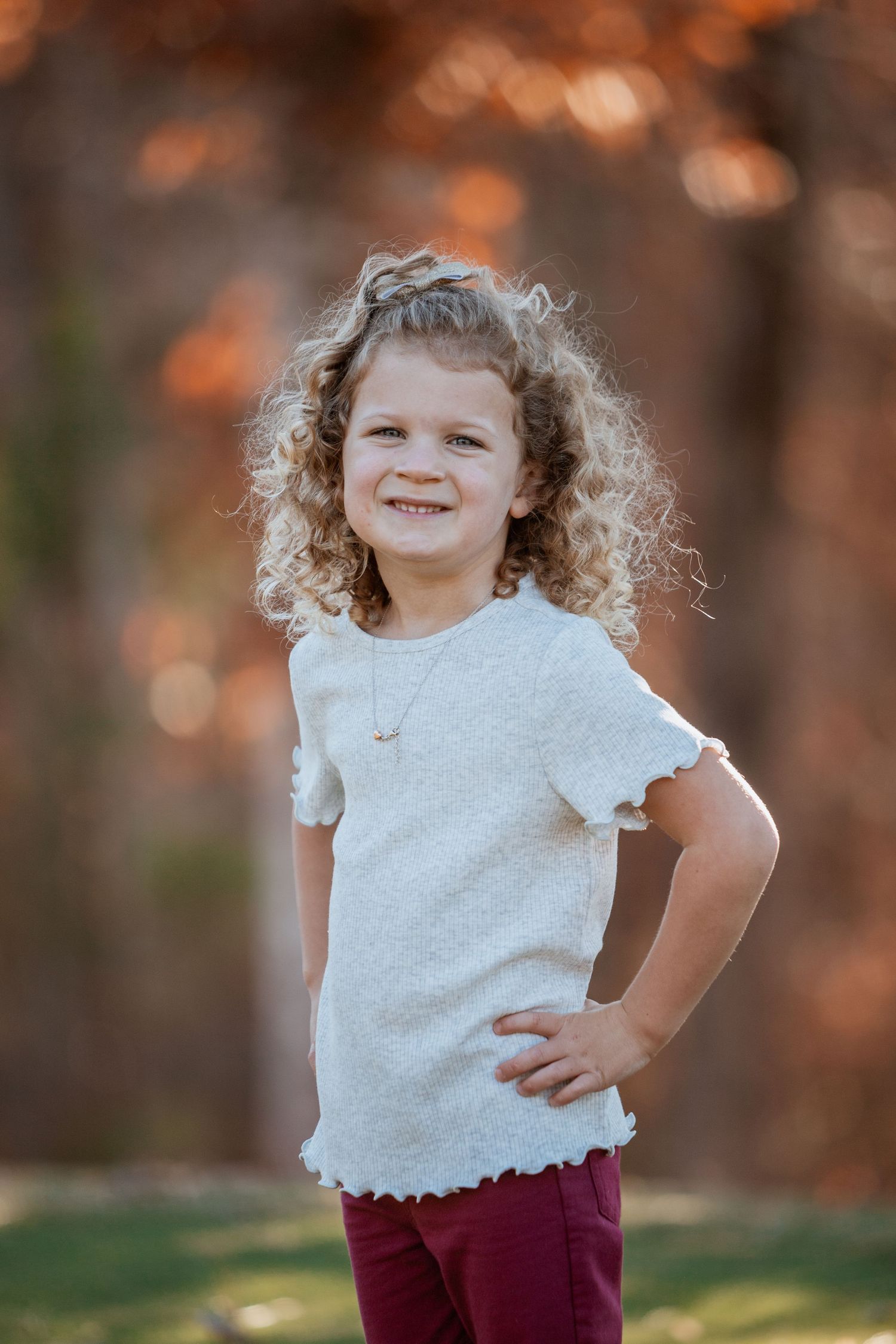 A cheerful young girl with curly hair poses confidently in a white top and burgundy pants outdoors.