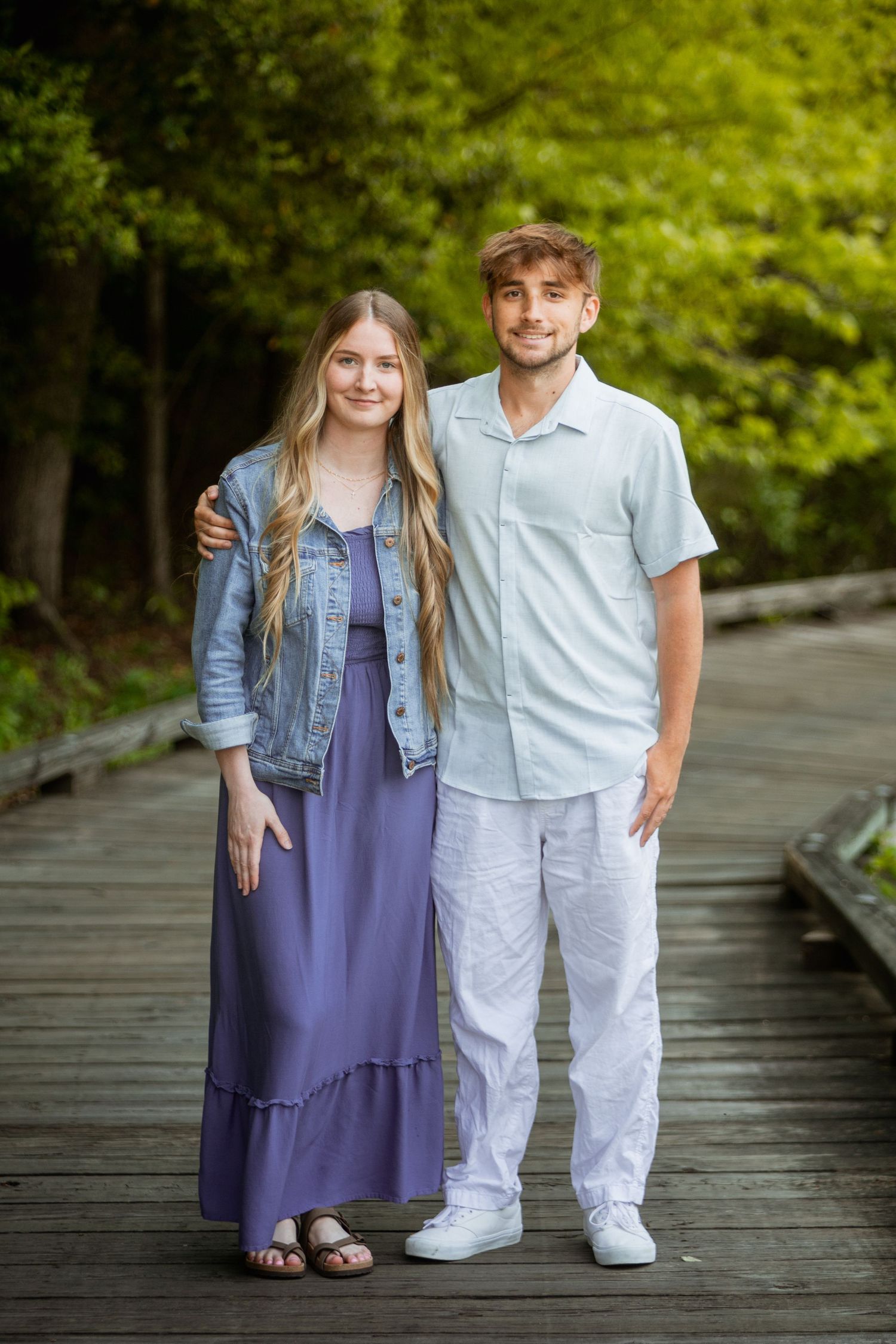 Young couple smiling together on a wooden boardwalk, woman in purple maxi dress and denim jacket.